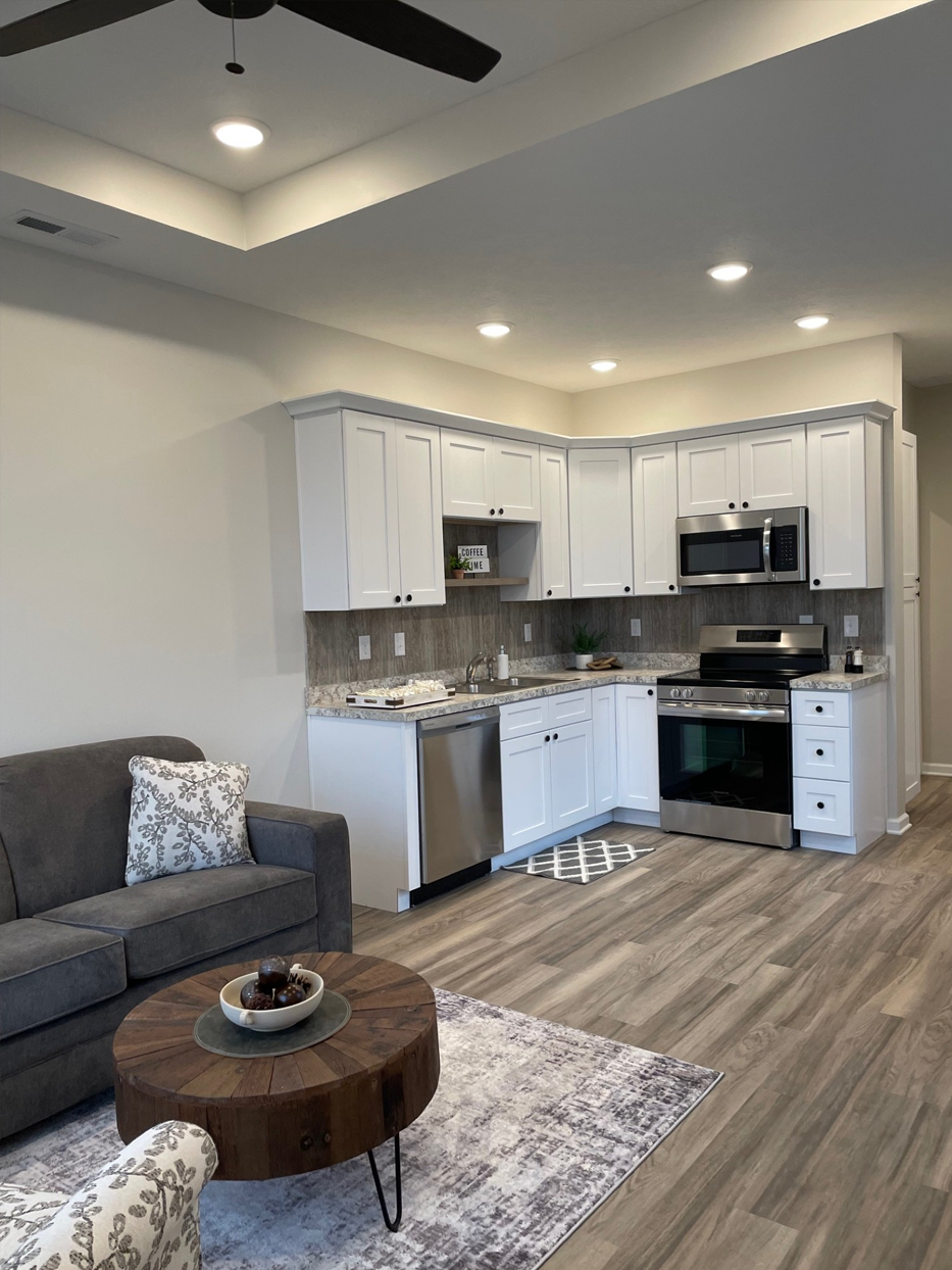 A modern kitchen with white cabinets, stainless steel appliances, and a living room with gray furniture and a wooden coffee table.