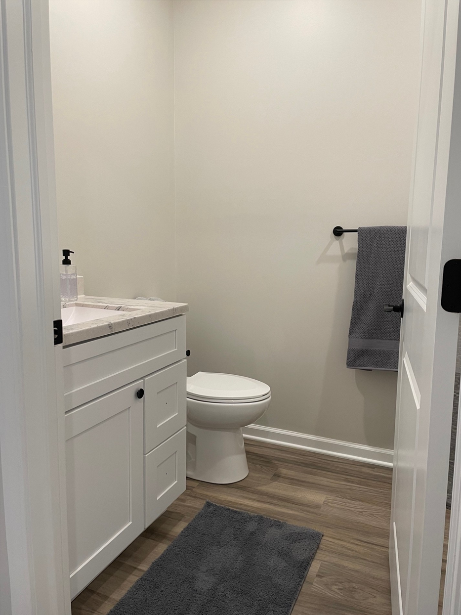 Small white bathroom with sink, toilet, and towel rack; gray rug and wood-look flooring.