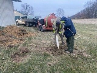 Two men are digging a hole in a field with a truck in the background.