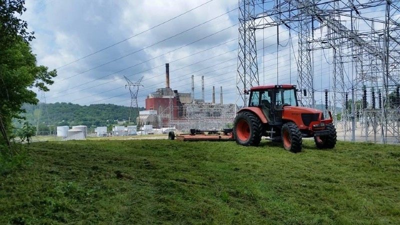 A red tractor is cutting grass in front of a power plant.