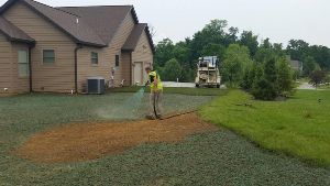 A man is spraying fertilizer on a gravel driveway in front of a house.