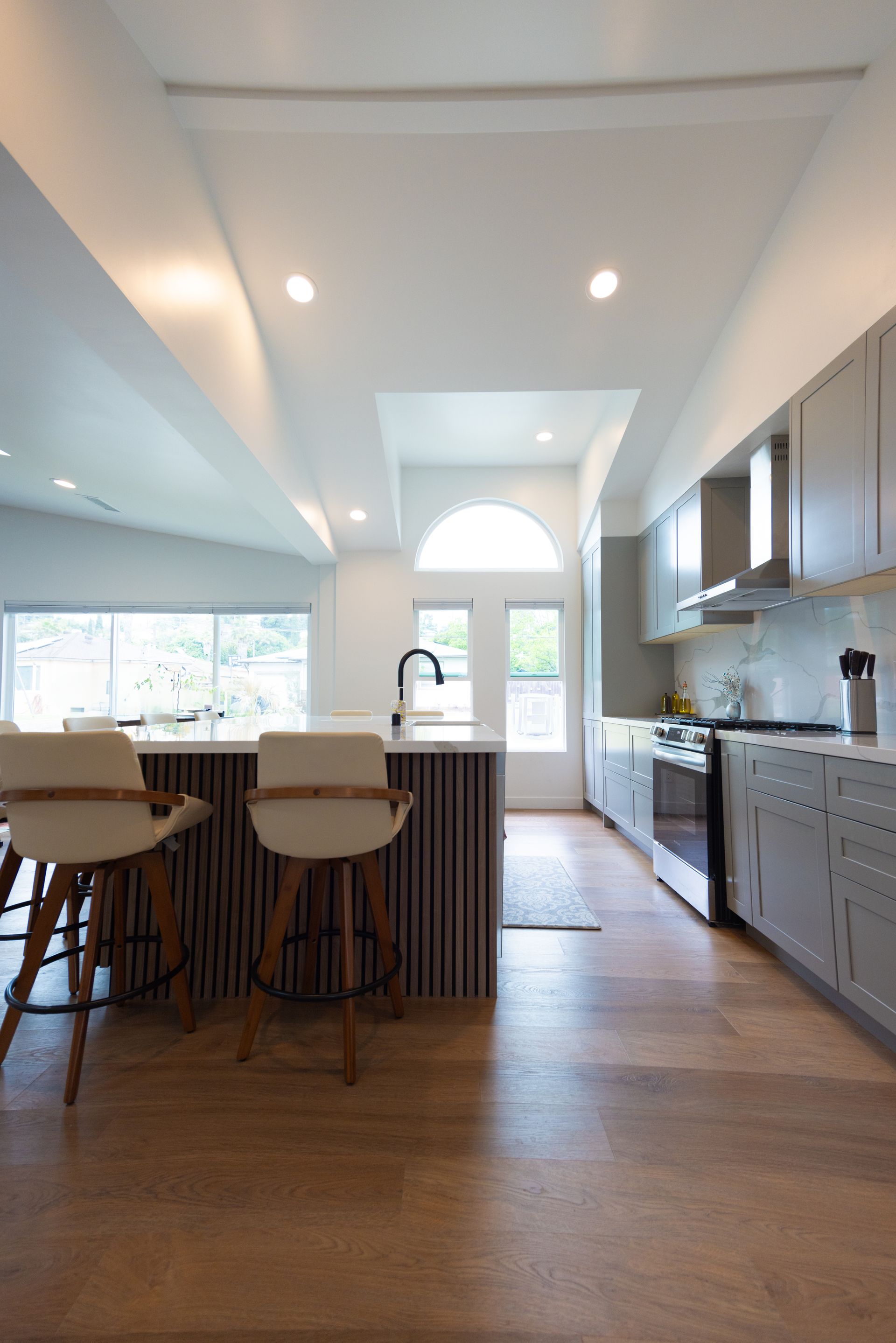 Modern kitchen with island, bar stools, wood floors, and bright windows leading to a back door.