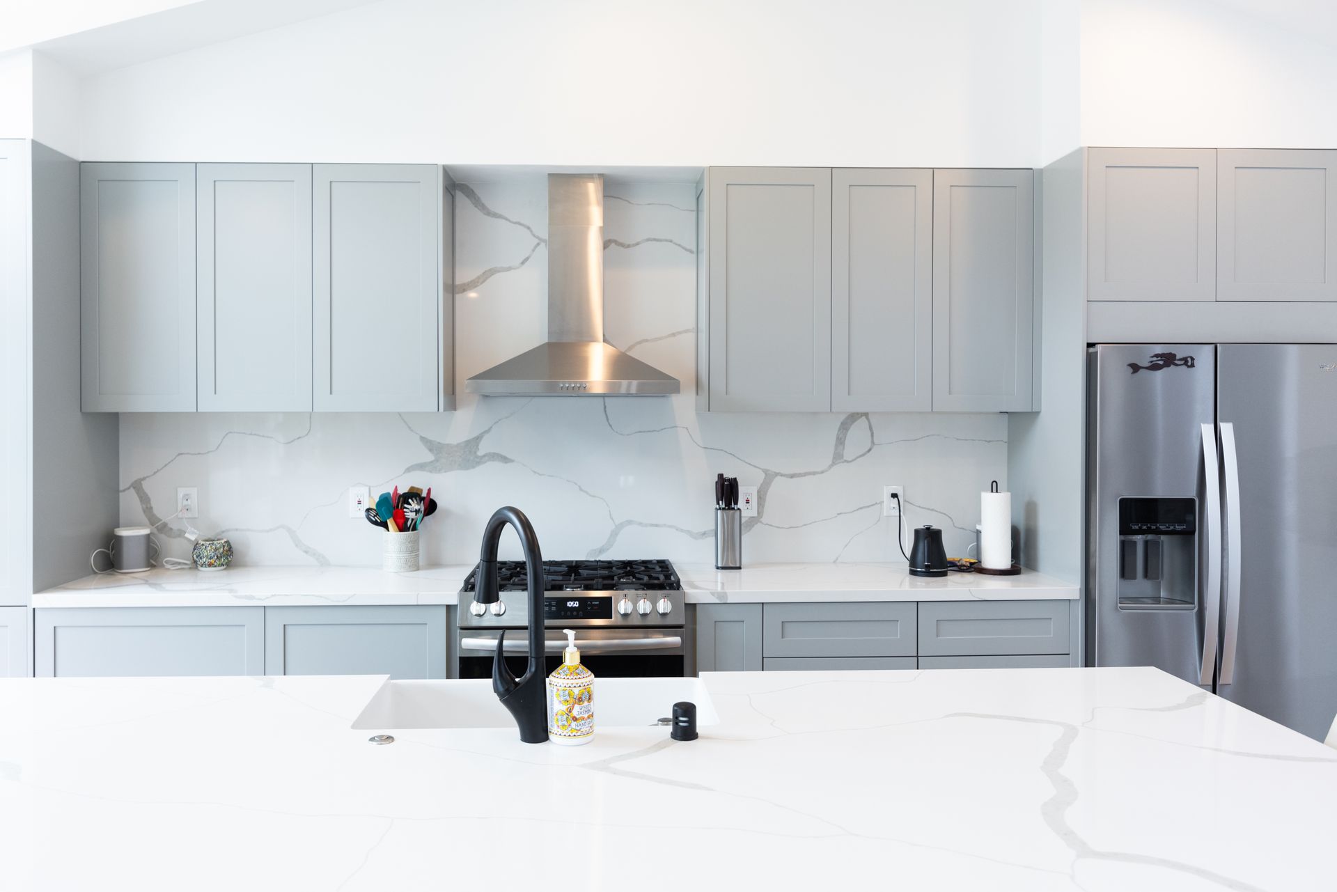 Bright white modern kitchen with marble island, black faucet, stainless appliances, and gray cabinets