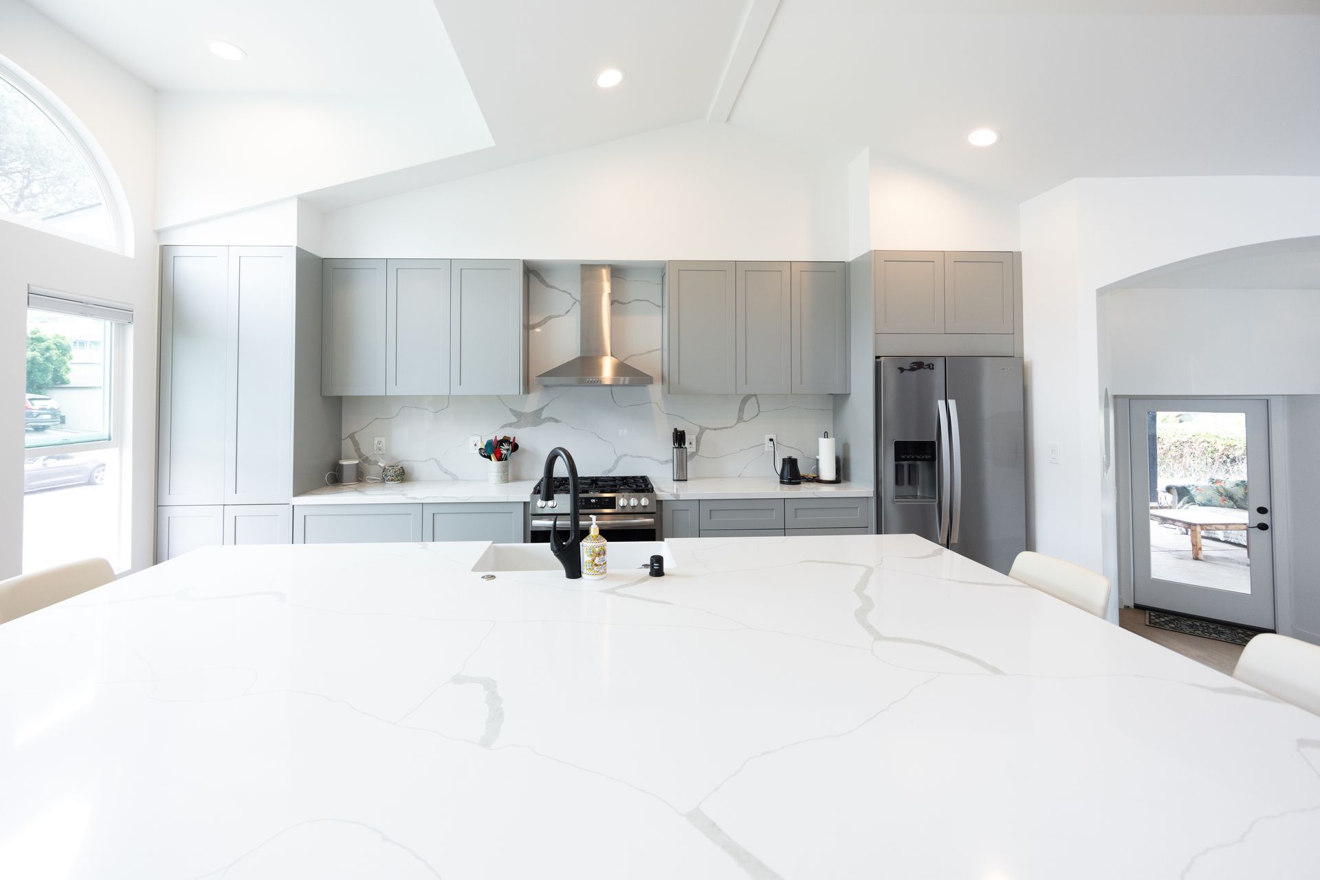 Bright modern kitchen with white island, gray cabinets, stainless range hood, and large windows