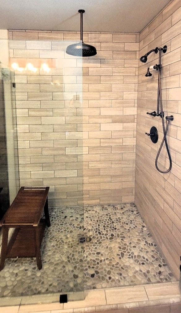 Modern shower with beige tile walls, pebble floor, glass door, bench, and black rainfall showerhead.
