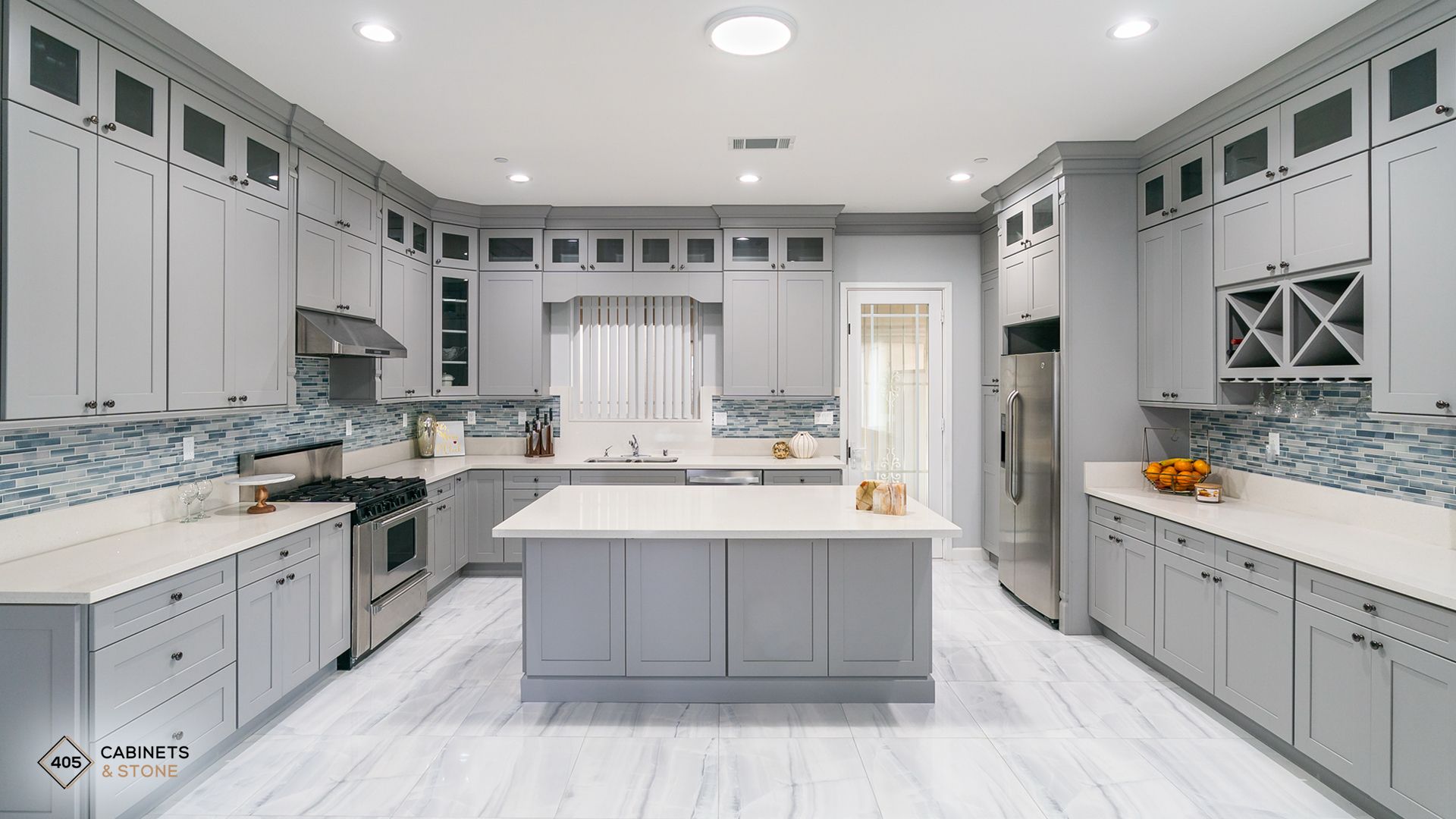 Bright modern kitchen with gray cabinets, white island, marble counters, and recessed ceiling lights.