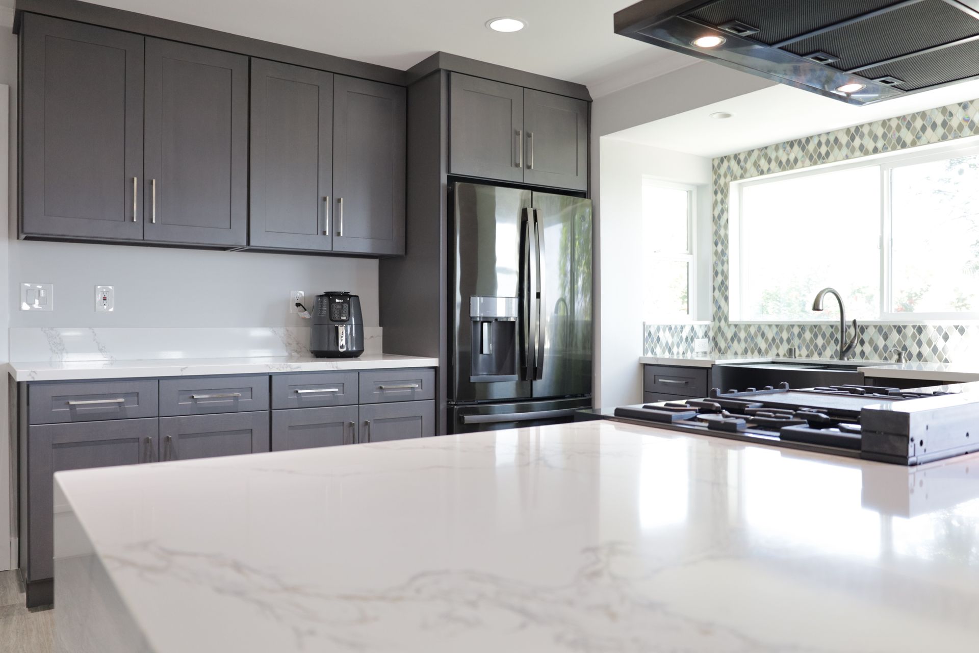 Modern kitchen with gray cabinets, black fridge, marble island, and large window over stove
