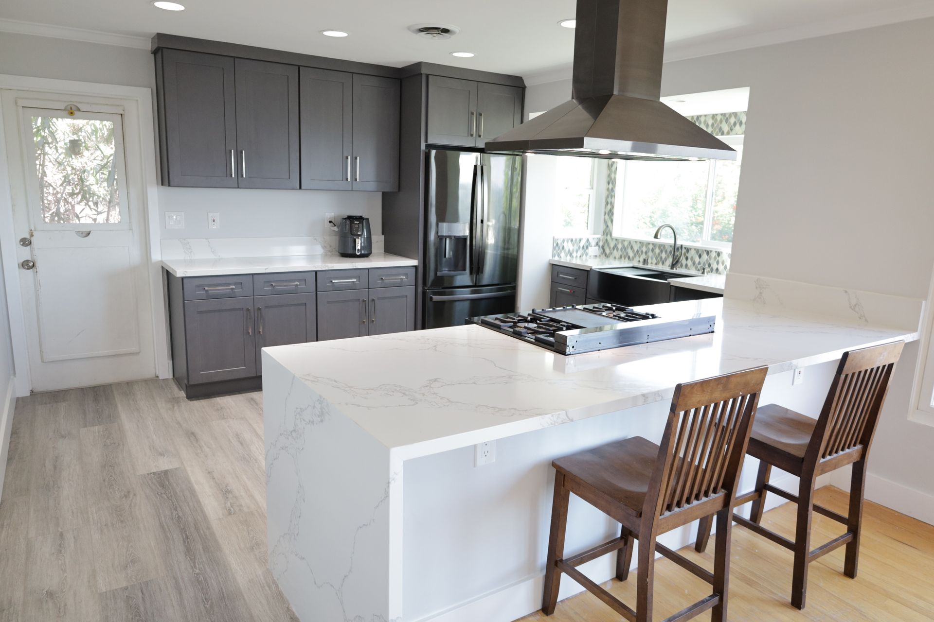 Modern kitchen with gray cabinets, white island, stainless range hood, and two wooden stools