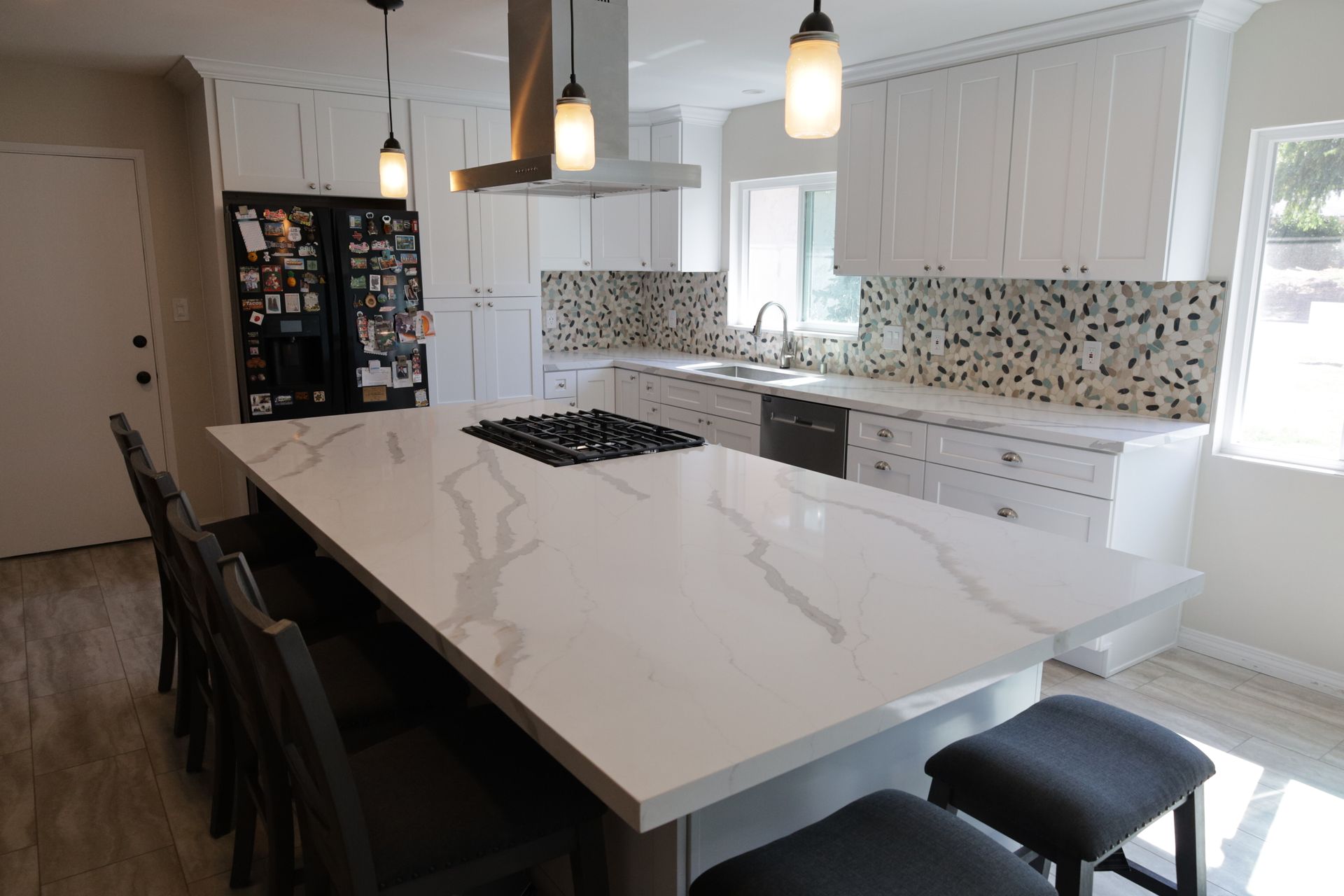 Modern kitchen with a white island, pendant lights, dark chairs, mosaic backsplash, and a bright window