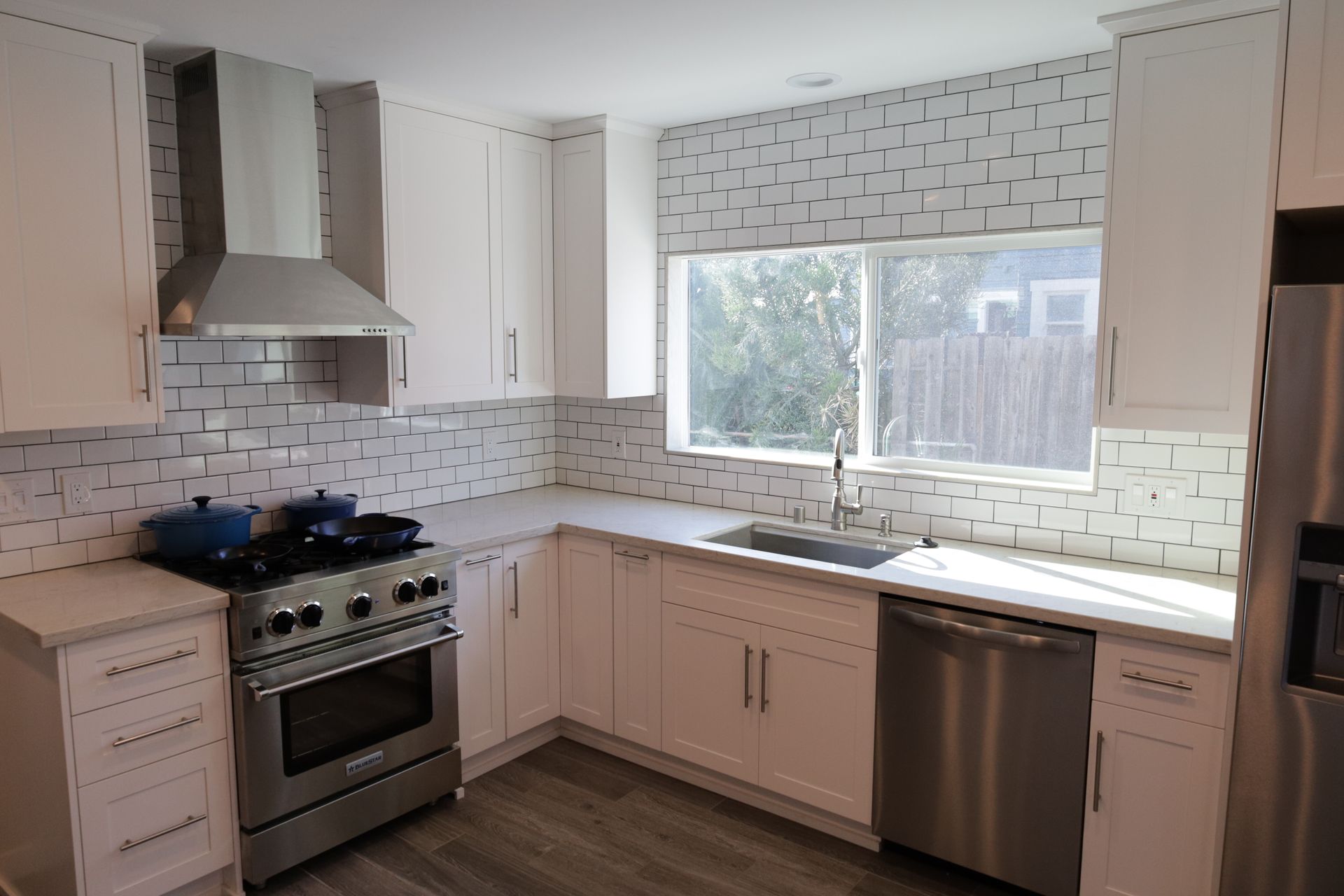 Modern white kitchen with stainless stove, tiled backsplash, large window, and dark wood floor