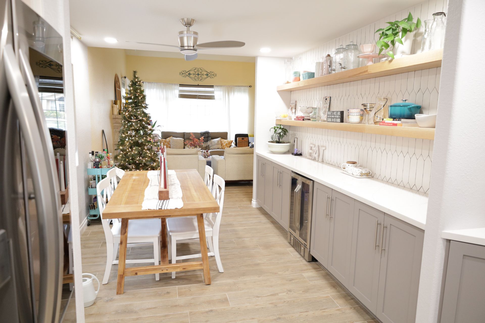 Bright kitchen with long dining table, gray cabinets, shelves, and a decorated Christmas tree.