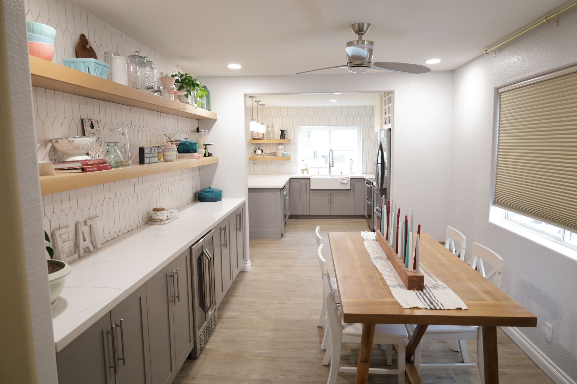 Bright kitchen dining area with long wooden table, gray cabinets, open shelves, and sunlight through windows