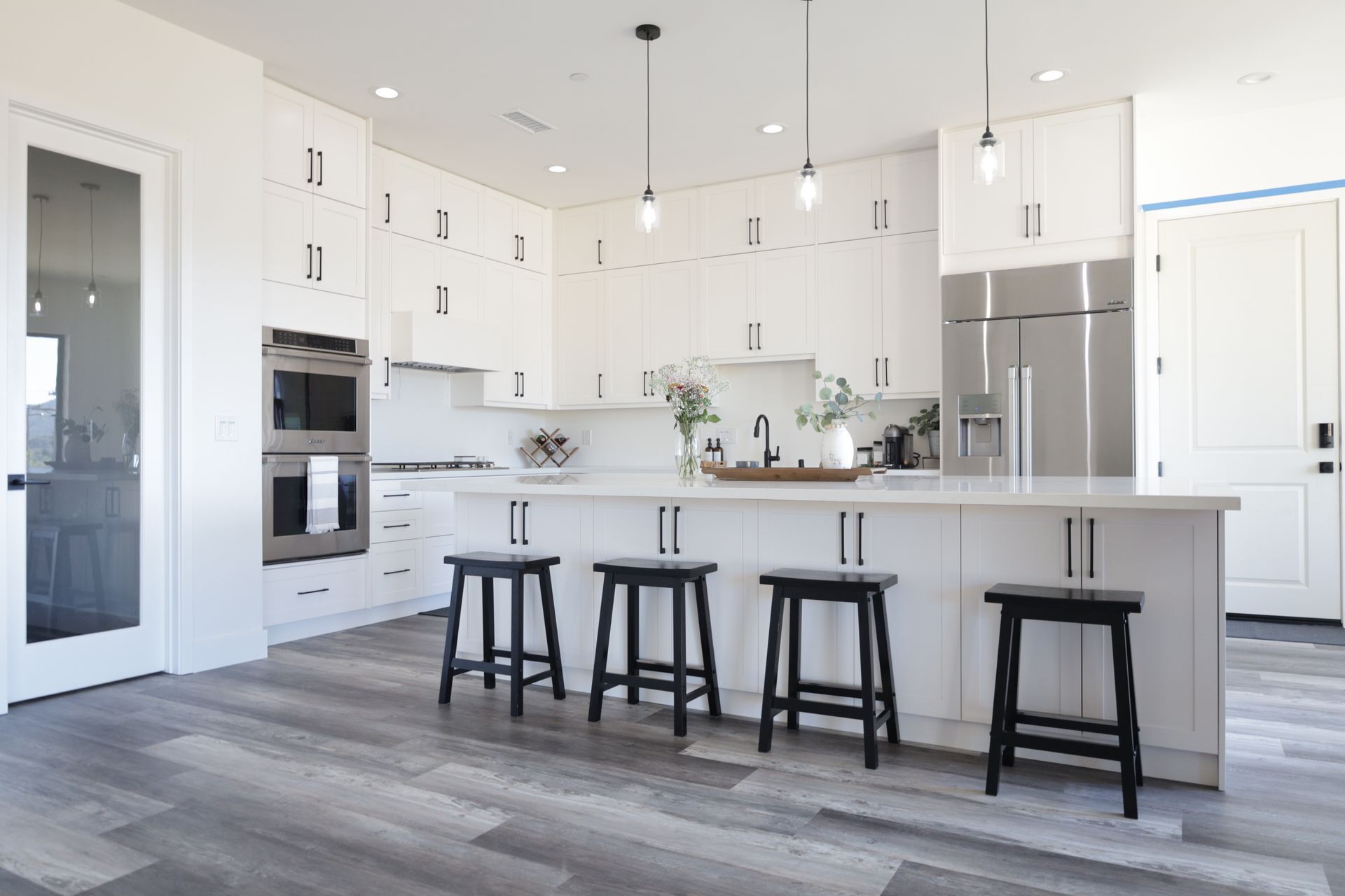 Bright modern white kitchen with island, black stools, stainless appliances, and gray wood floors