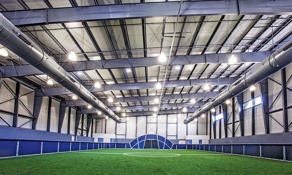 Indoor soccer field with green turf, blue walls, and metal rafters under bright lights