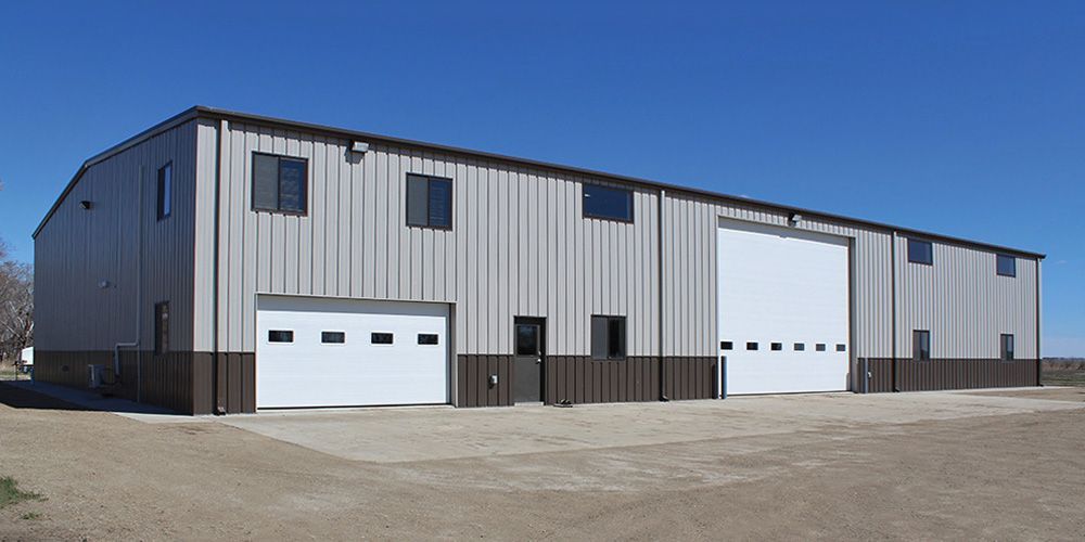 Large gray industrial warehouse with white garage doors on a gravel lot under a clear sky