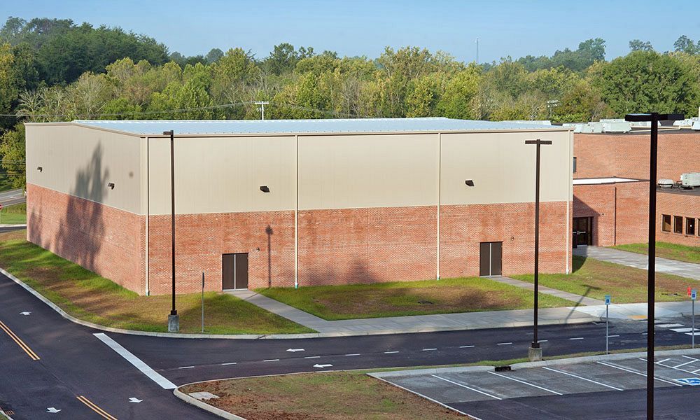 Large brick-and-beige industrial building beside a road and parking area, with trees in the background.