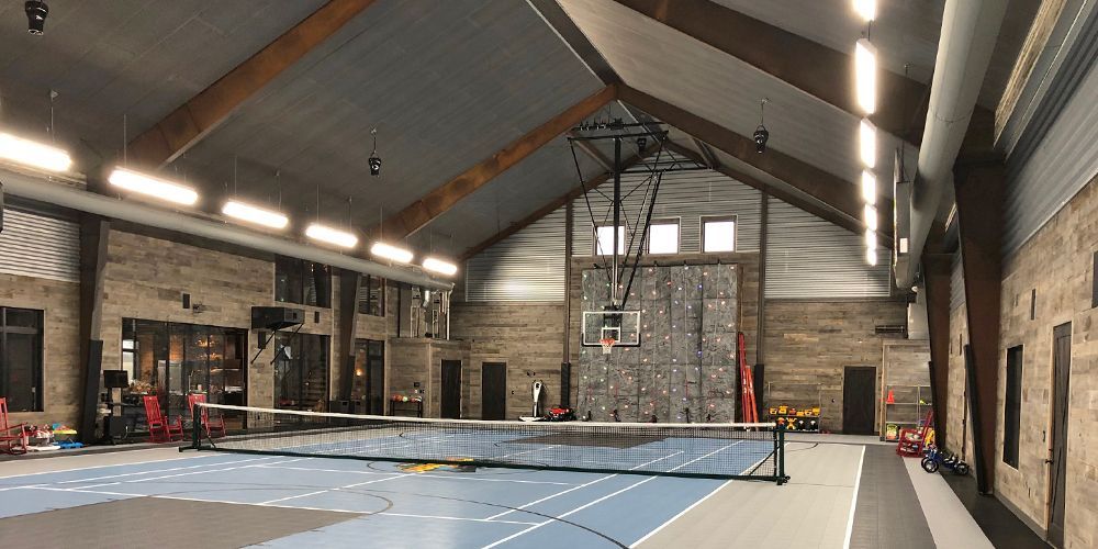 Indoor pickleball court in a rustic hall with nets, tall windows, and high exposed wood beams