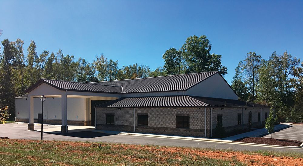 Single-story brick building with a dark metal roof, white columns, and a paved driveway surrounded by trees.