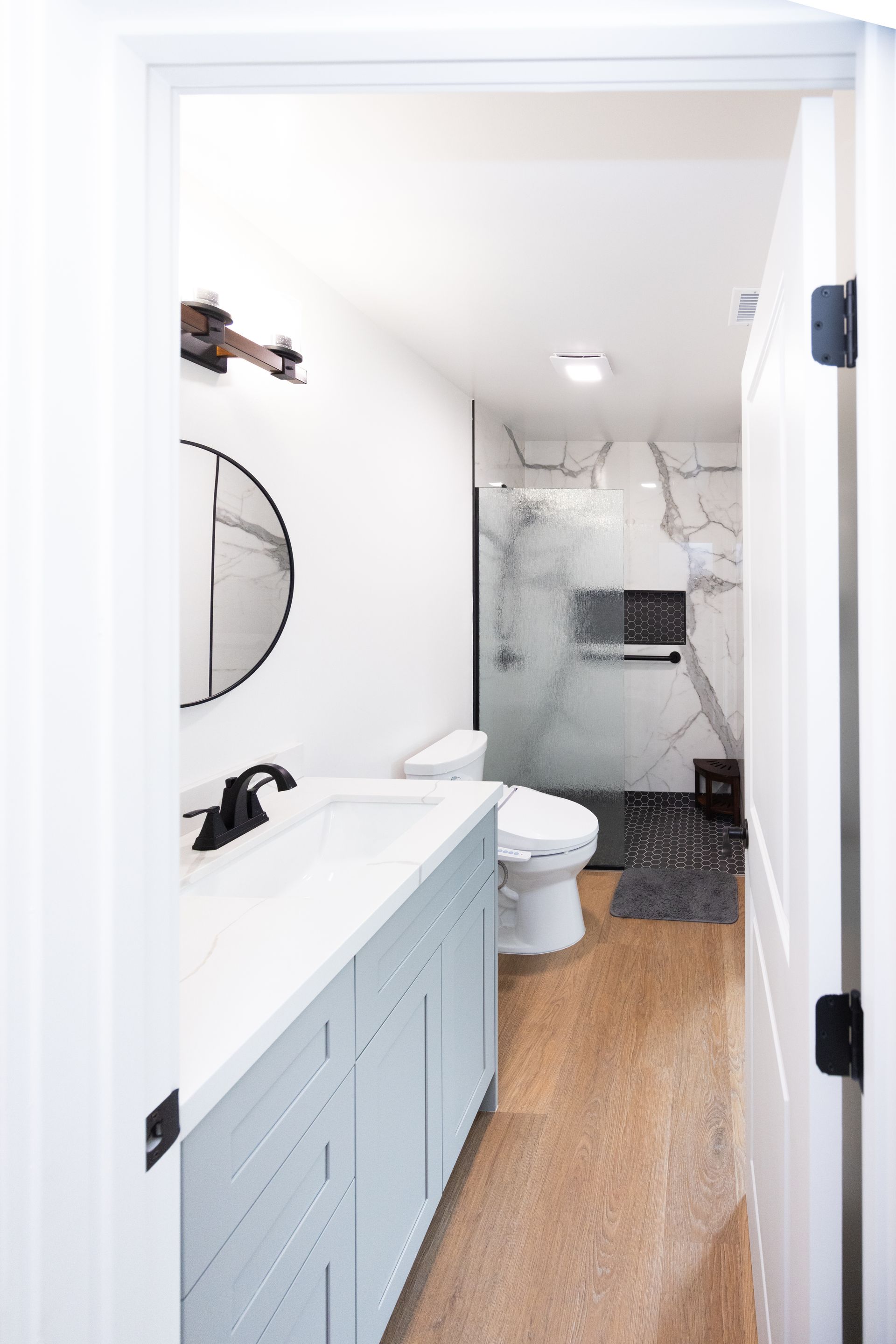 Small bathroom with blue vanity, toilet, wood floor, and a tiled shower at the back.