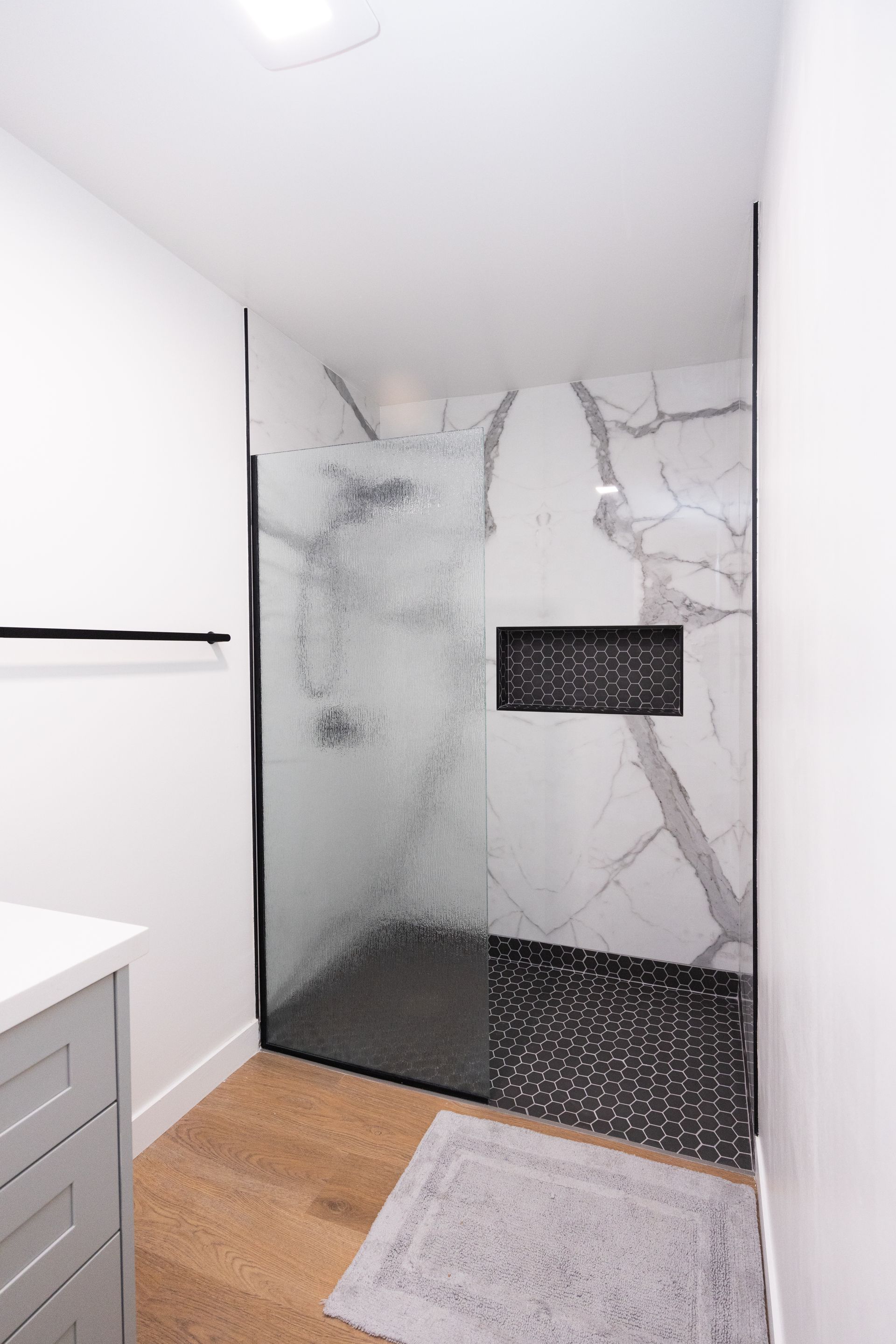 Modern shower with marble-look walls, black hex tile floor, and gray bath mat in a narrow bathroom.