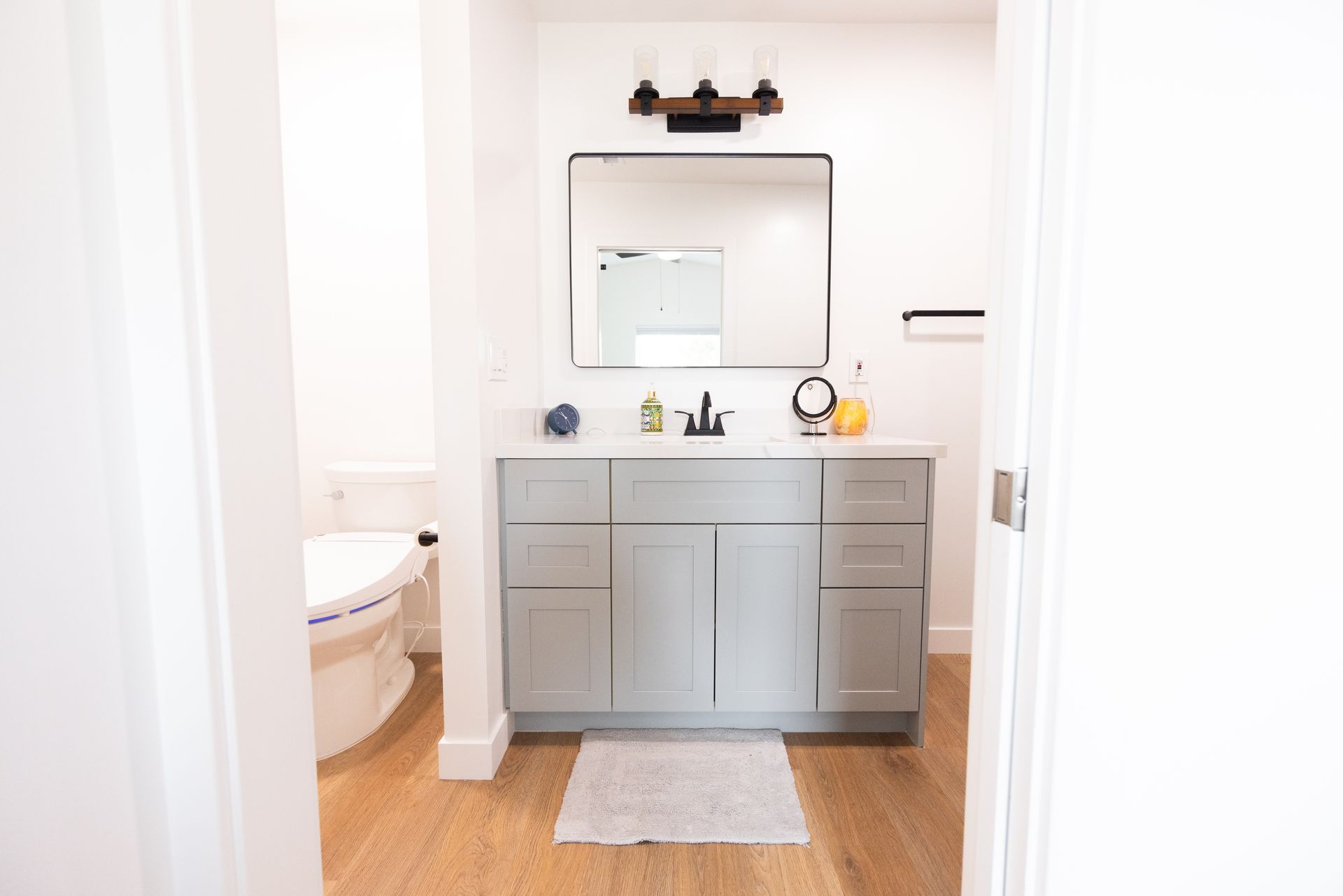 Bright bathroom vanity with gray cabinets, mirror, sink, tub, and wood floor viewed through a doorway