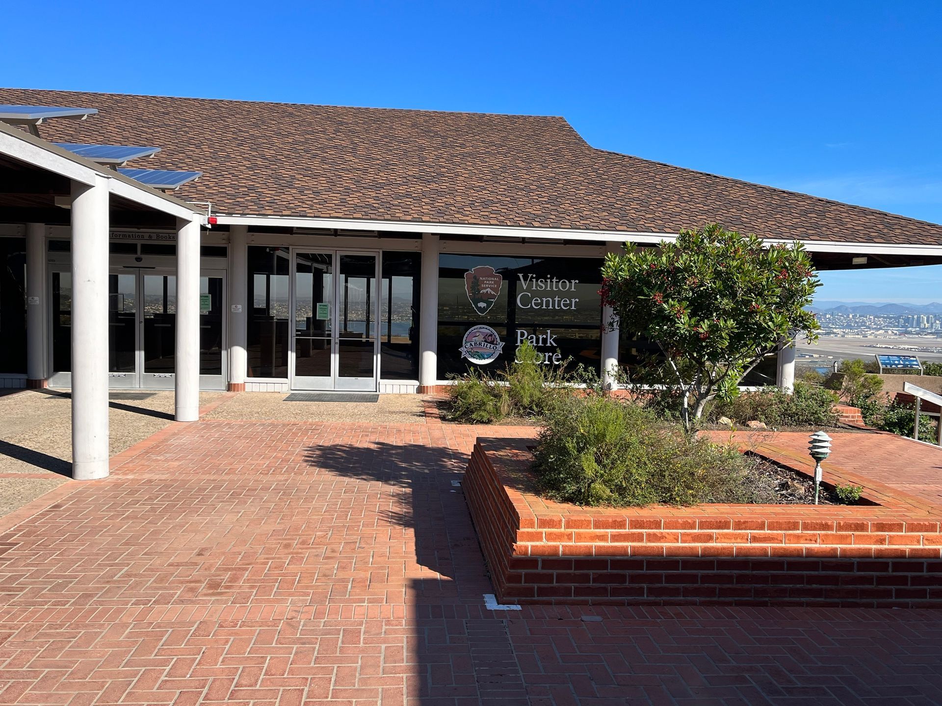 Brick courtyard outside a storefront with glass doors, red-tile roof, and ocean view in the distance