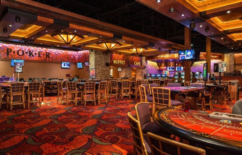 Empty casino floor with poker tables, red patterned carpet, wooden chairs, and warm ambient lighting