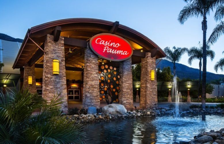 Casino entrance at dusk with a red sign, stone columns, palm trees, and a reflecting water fountain