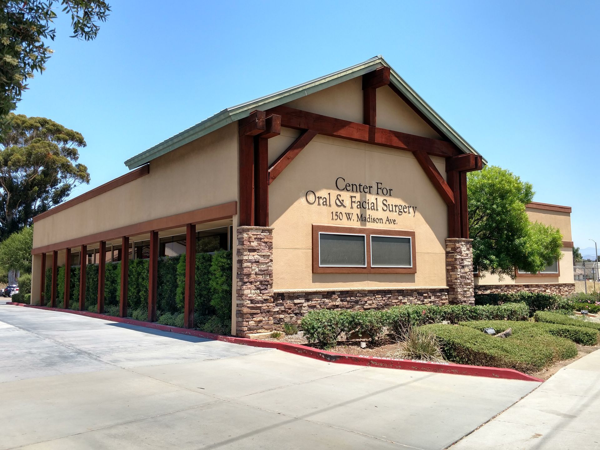 Exterior of a tan community center building with a sign, red trim, and landscaped shrubs under a clear blue sky