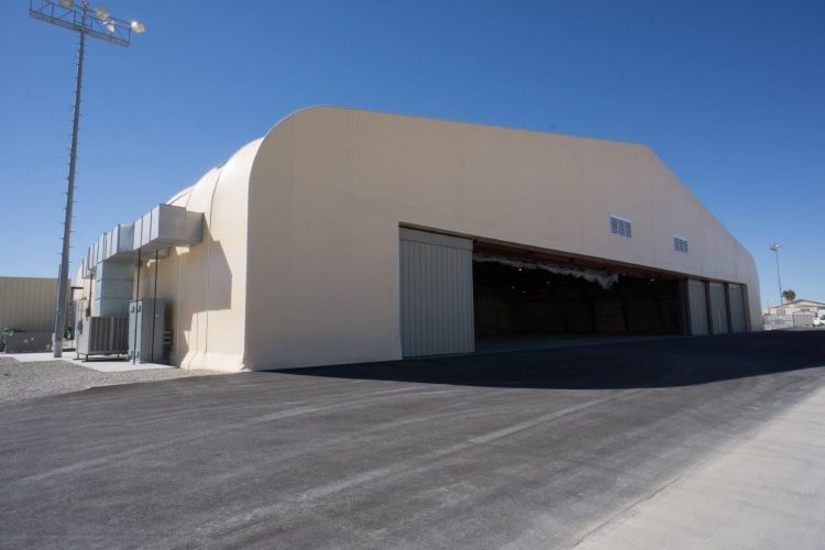 Large beige industrial hangar with open doorway on a paved lot under a clear blue sky
