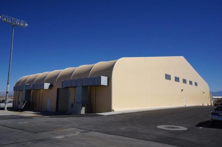 Large beige industrial building with a curved roof and small windows in an empty parking lot under a clear blue sky