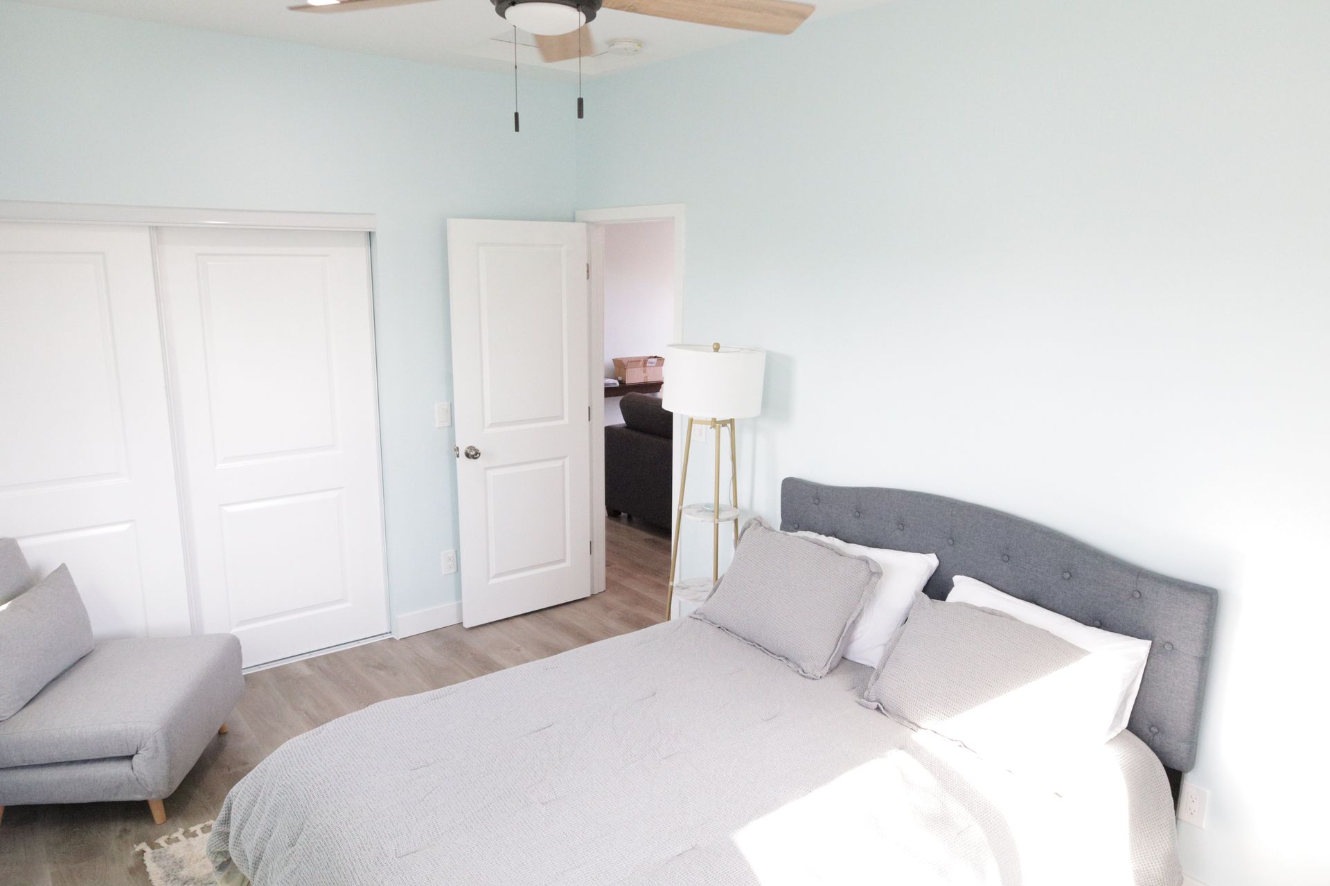 Bright bedroom with a bed, gray chair, white closet doors, and pale blue walls.