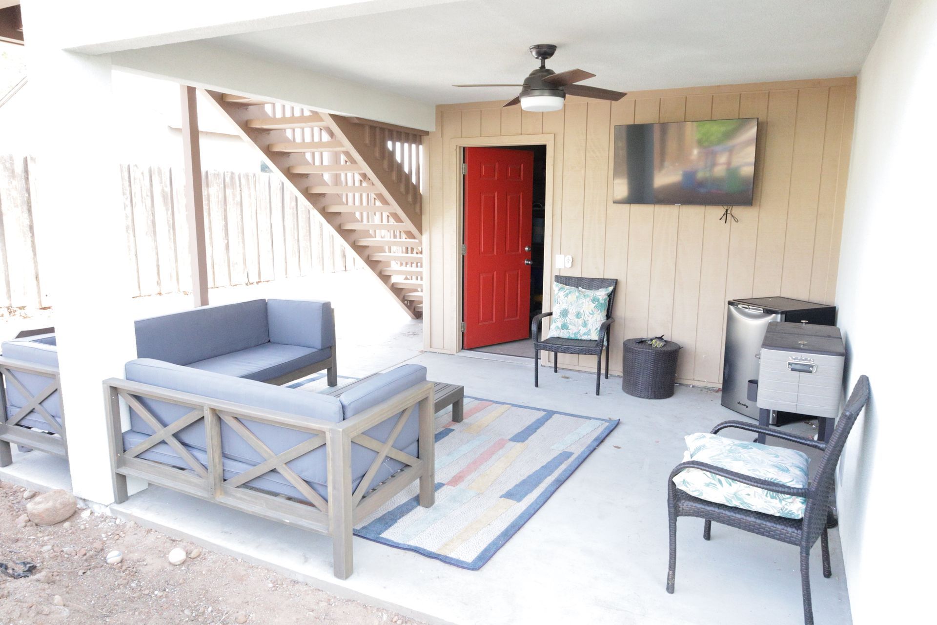 Covered patio with gray seating, red door, stairway, rug, and wall-mounted TV