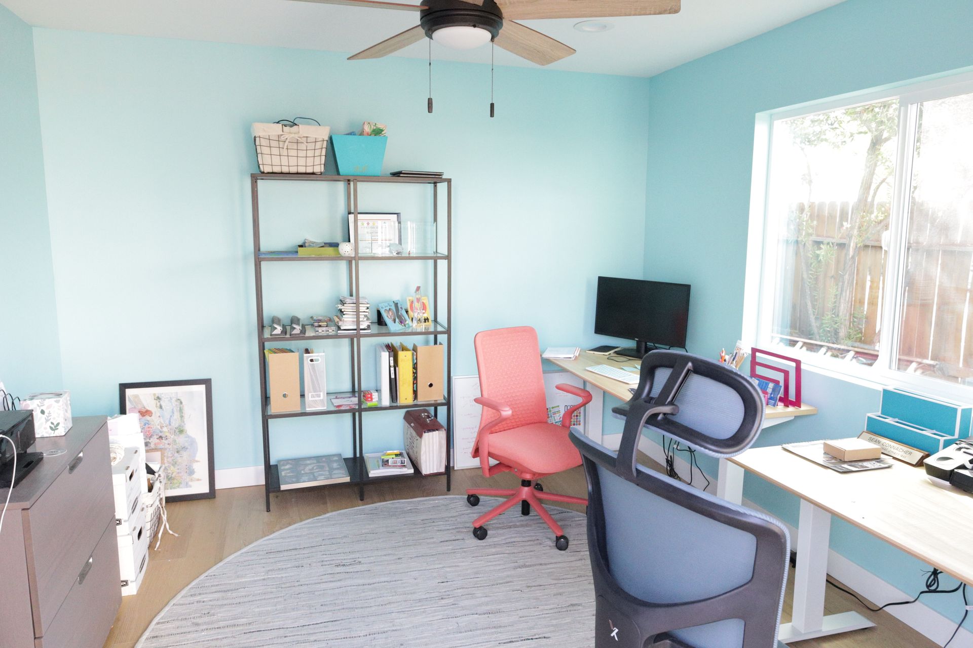 Bright home office with blue walls, desk, computer, shelves, and two chairs by a window