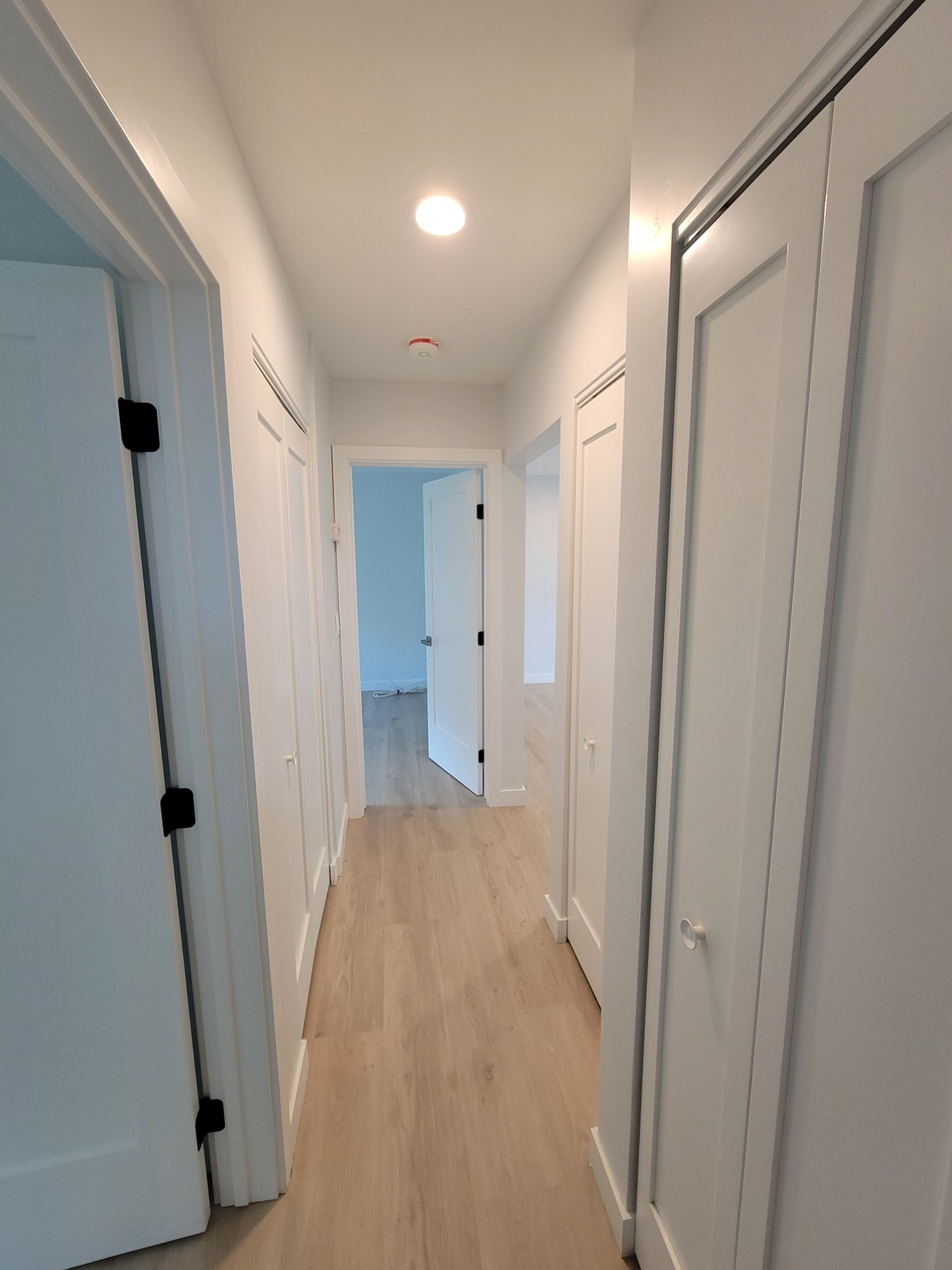 Bright hallway with white doors, recessed lights, and light wood flooring leading to a blue room.