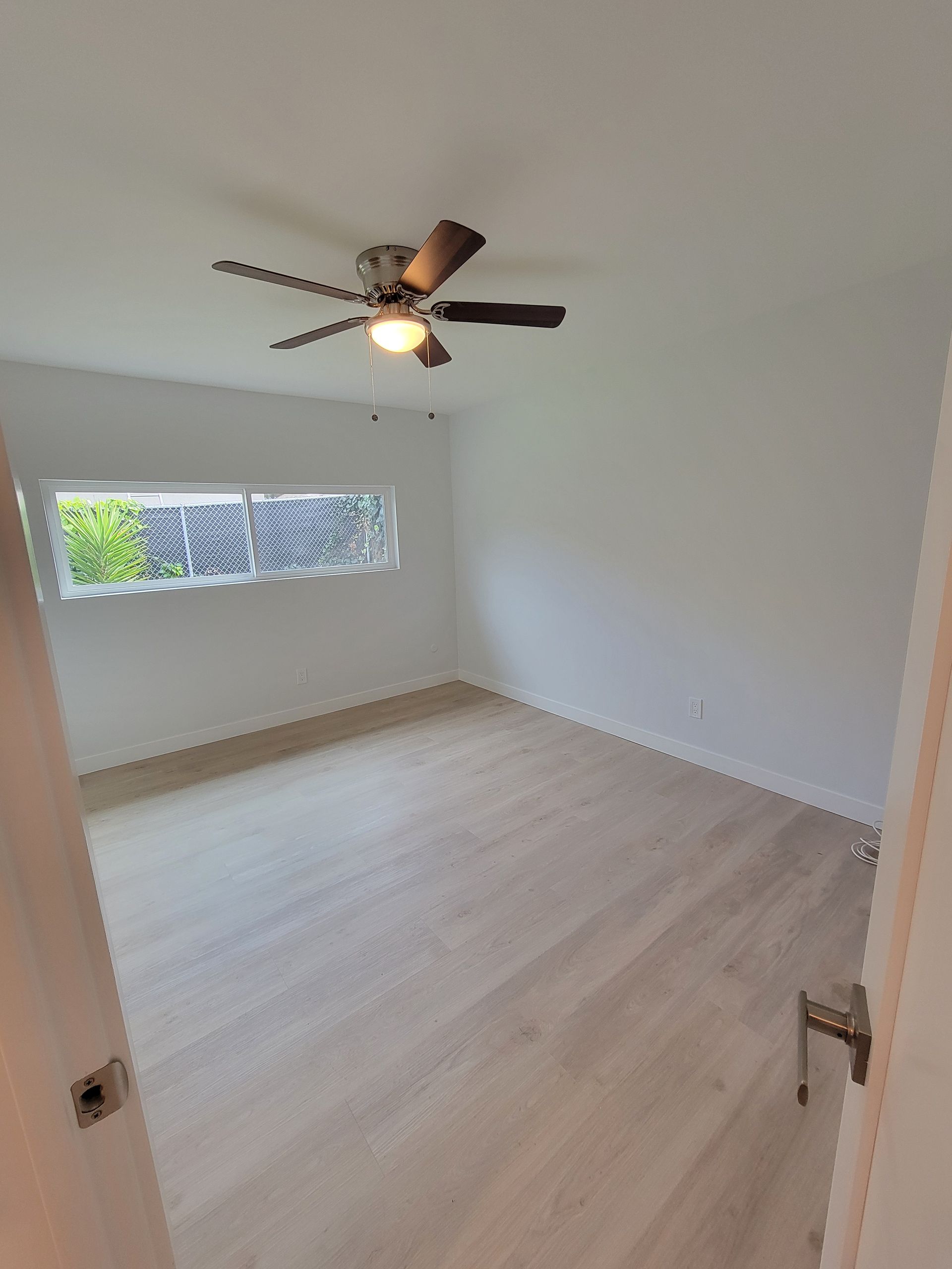 Empty white bedroom with light wood floor, ceiling fan, and a small window with greenery outside