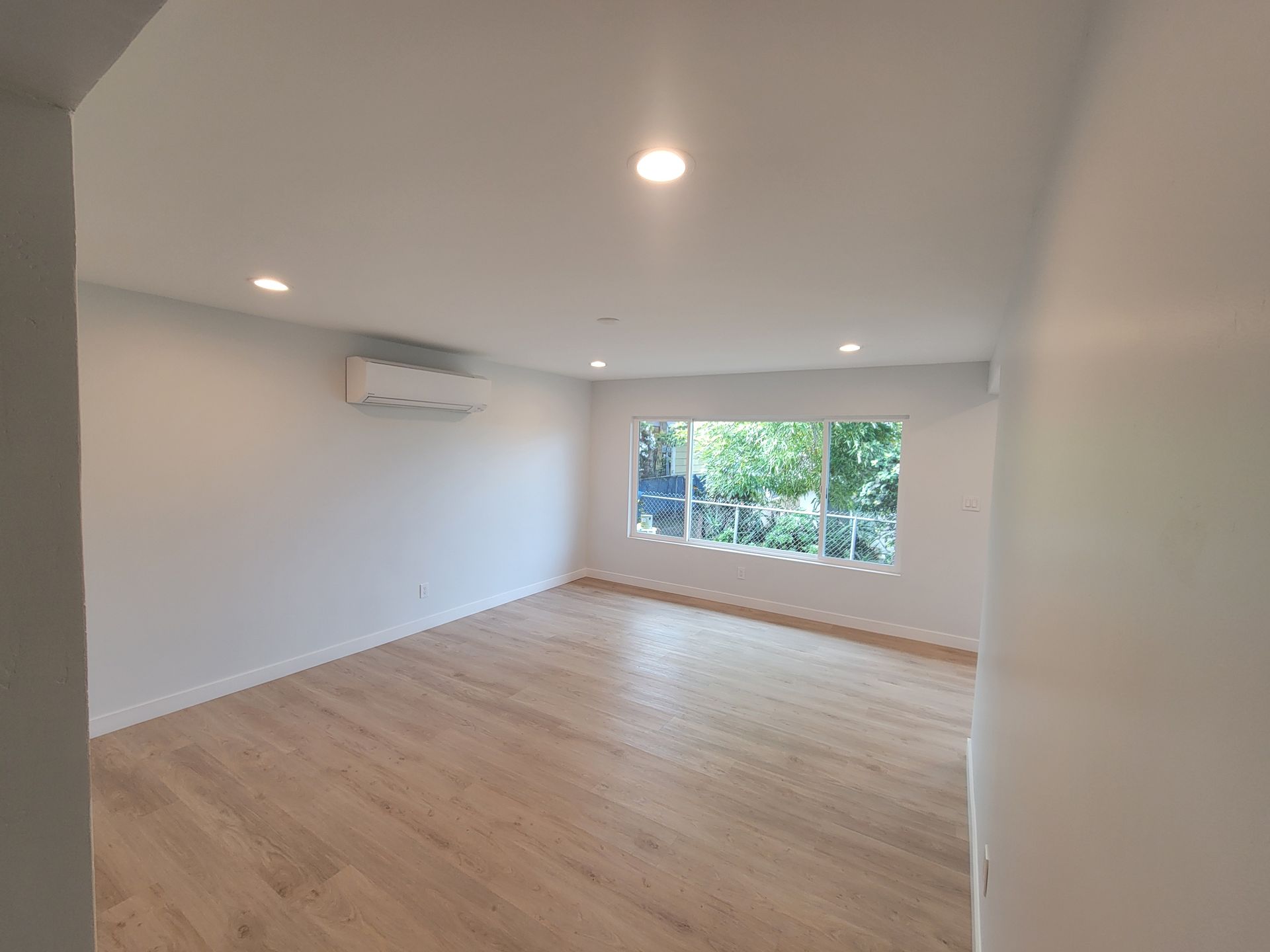 Empty bright living room with light wood floors, recessed lights, wall-mounted AC, and a large window overlooking greenery