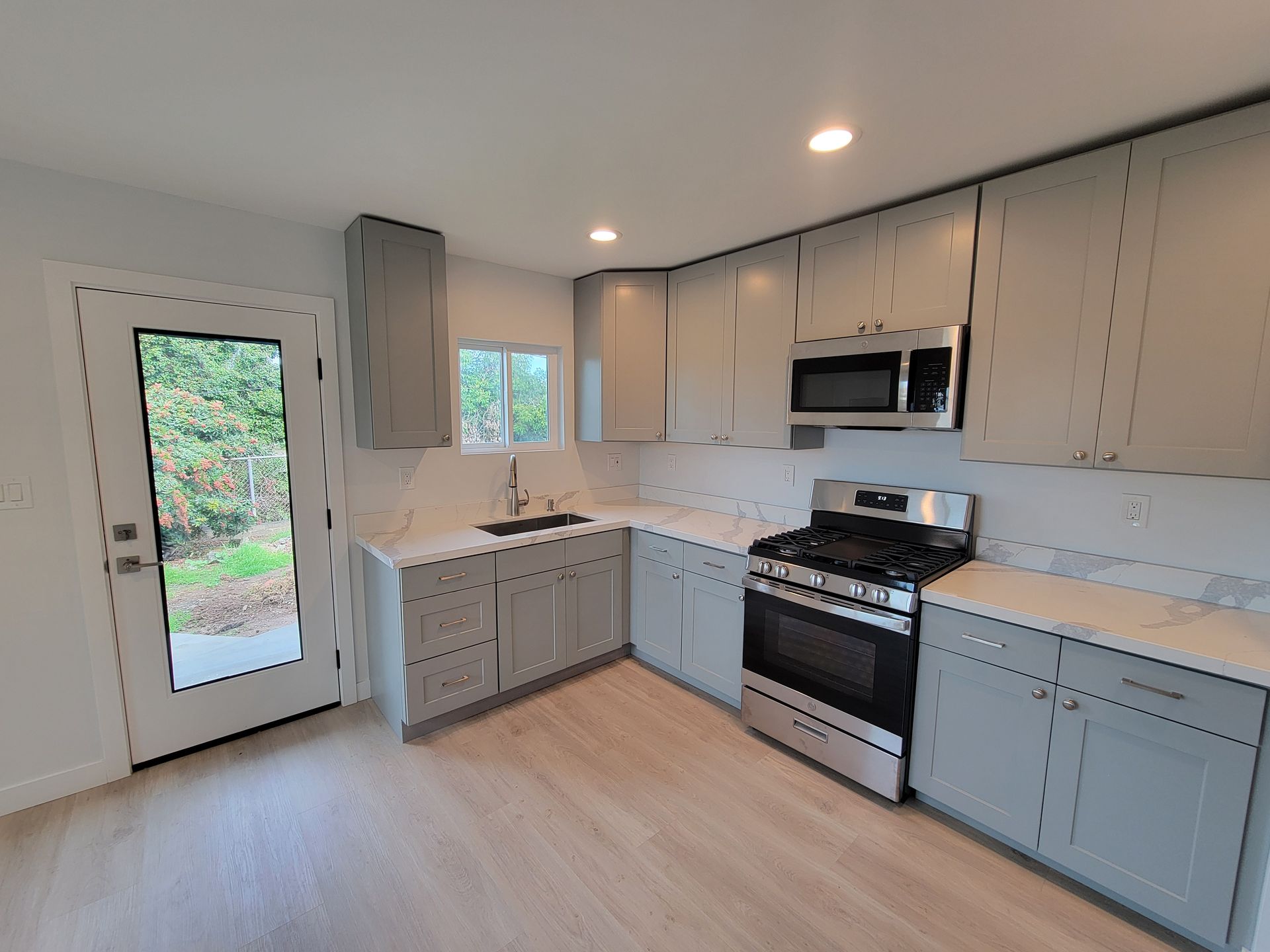 Modern kitchen with gray cabinets, white countertops, stainless appliances, and a door to greenery