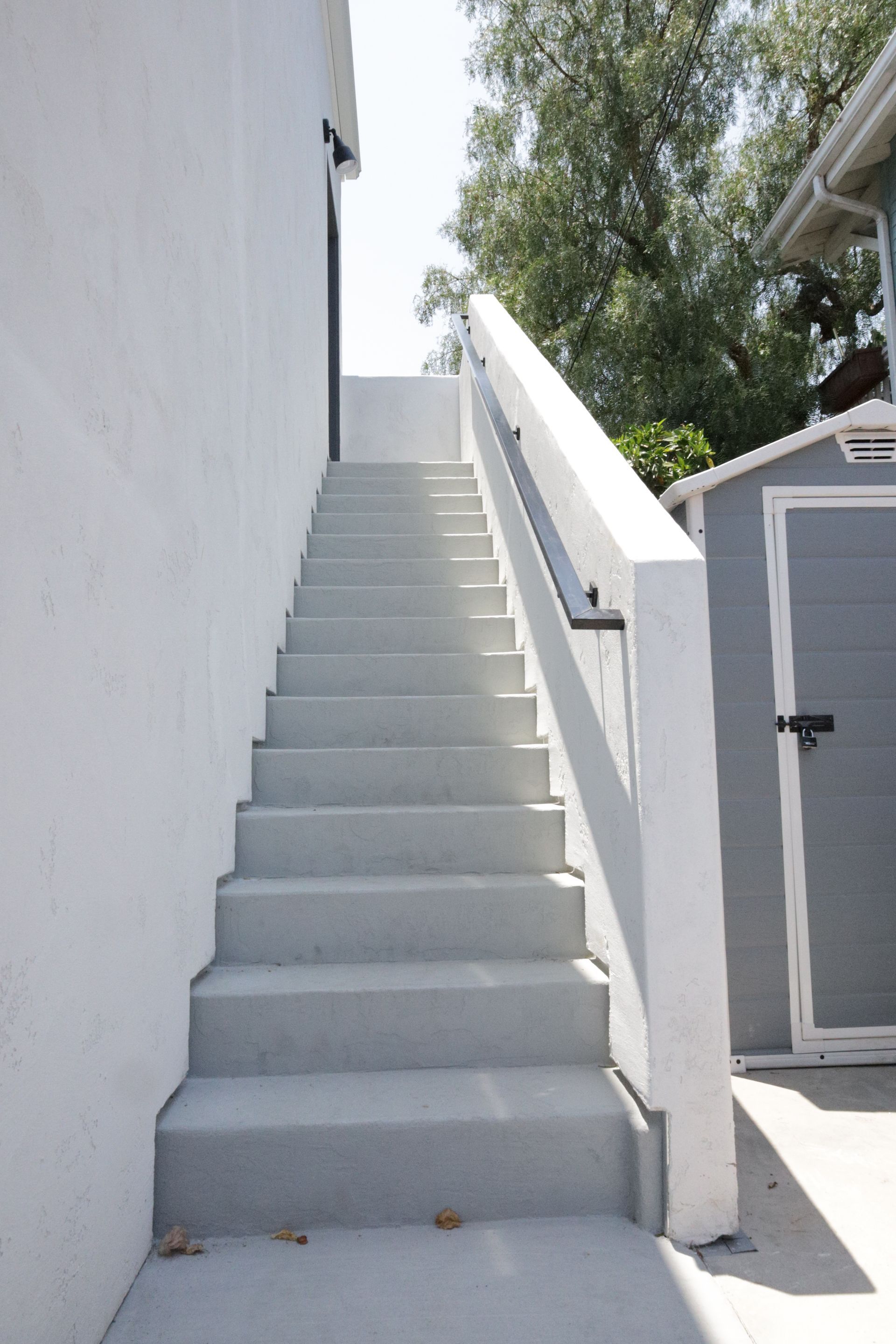 Steep outdoor concrete staircase beside a white wall, leading up to a gray garage and trees.