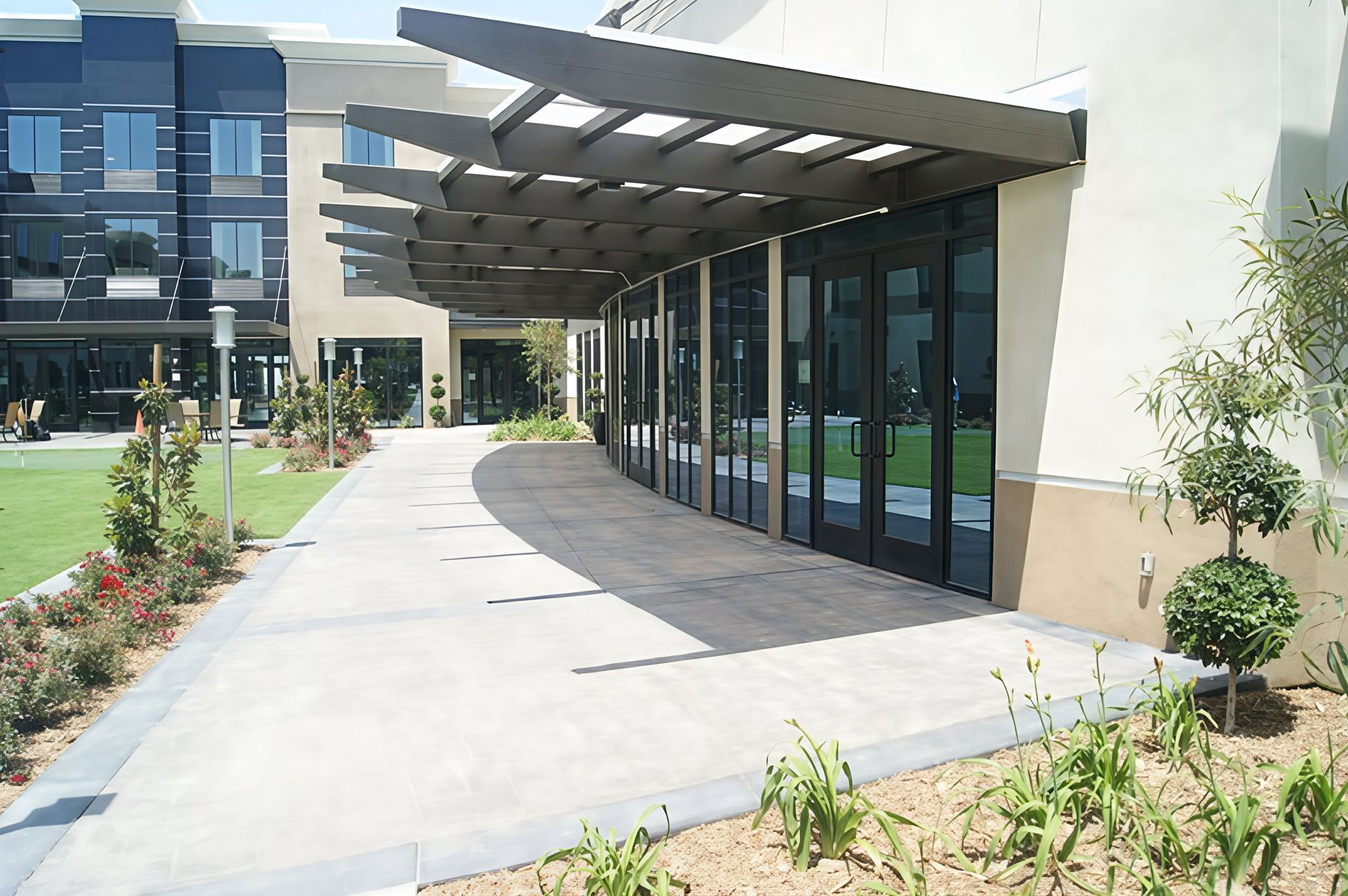 Modern building entrance with glass doors, covered walkway, and landscaped path in bright daylight