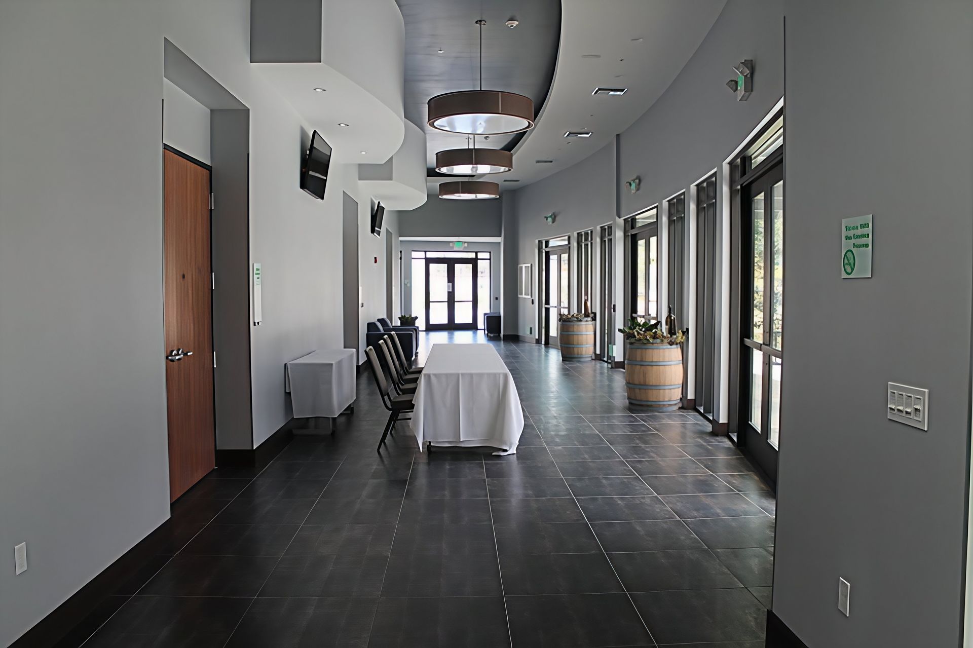 Long hallway with dark floor tiles, white walls, tables, chairs, and large windows along one side