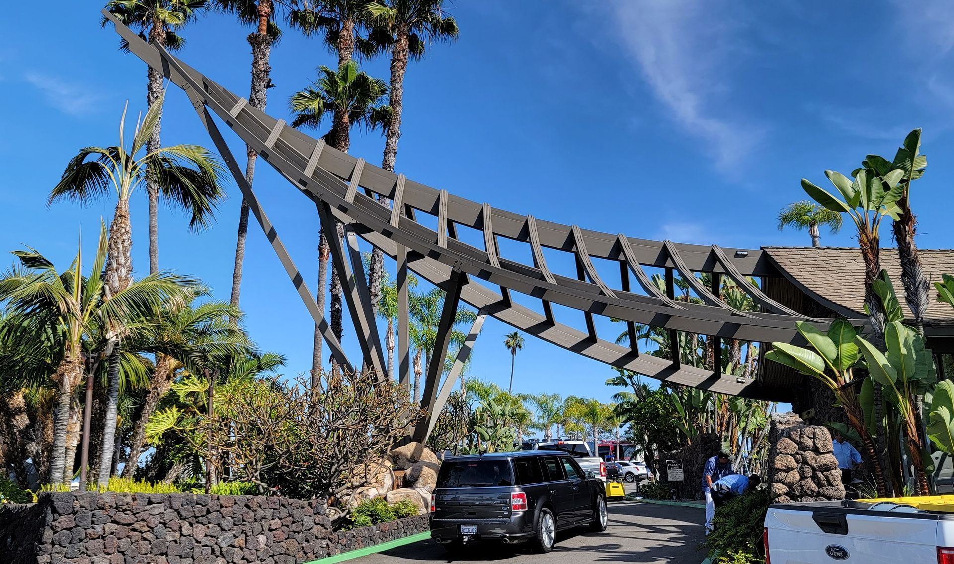 Suspended wooden bridge over tropical landscaping, with a black SUV on a road under a bright blue sky.