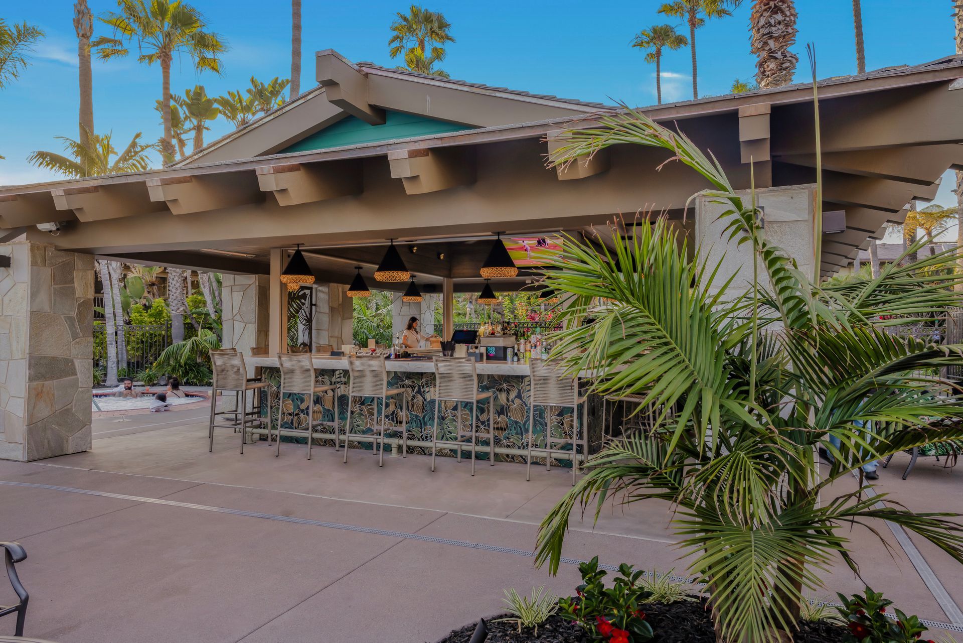 Open-air patio bar with stools, palm trees, and a tiled roof under a blue sky