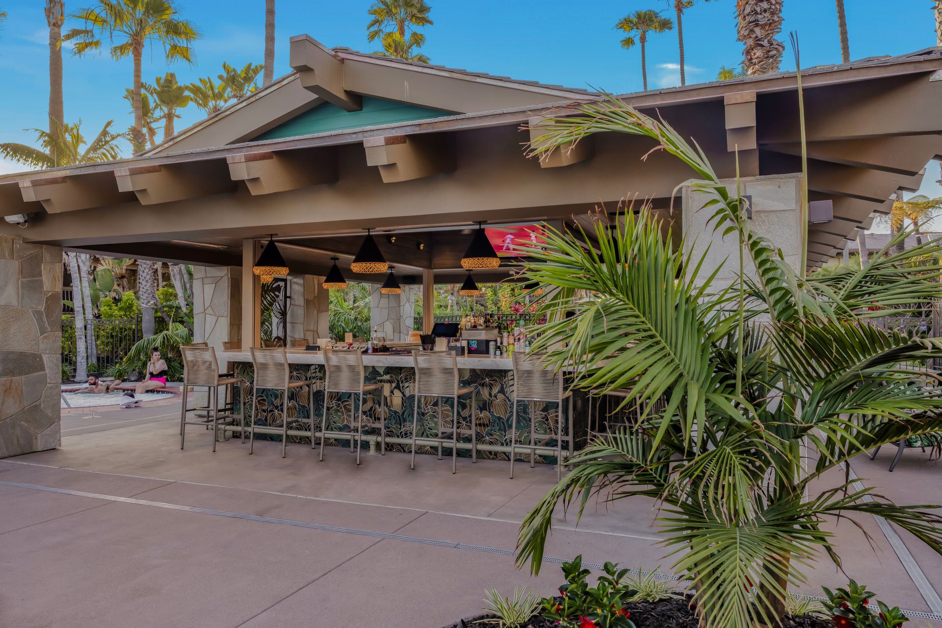 Outdoor restaurant patio with tables and chairs under a covered roof, surrounded by palm trees and greenery
