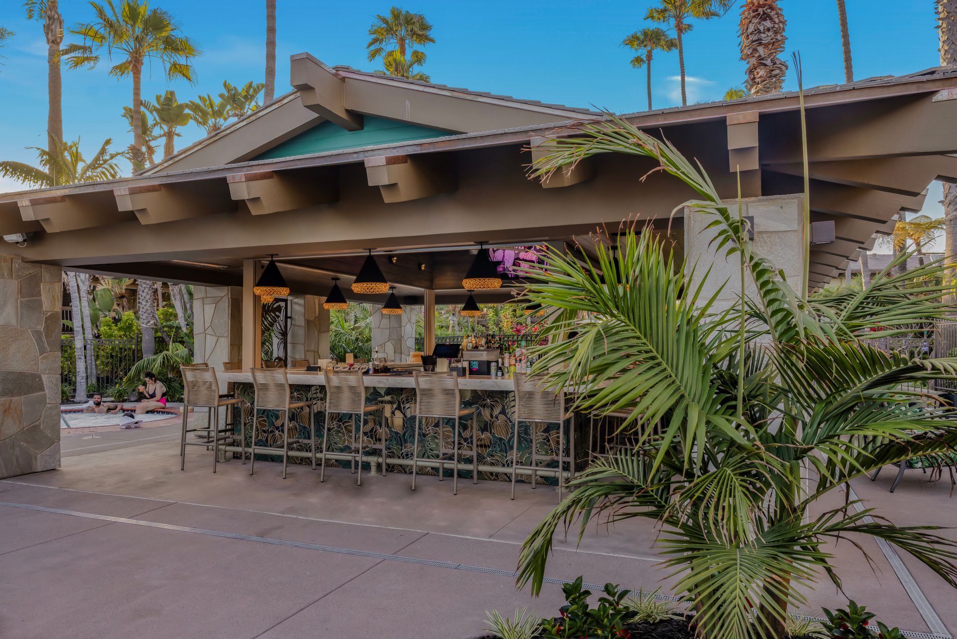 Outdoor patio with tables and chairs under a shaded roof, surrounded by palm trees and greenery.