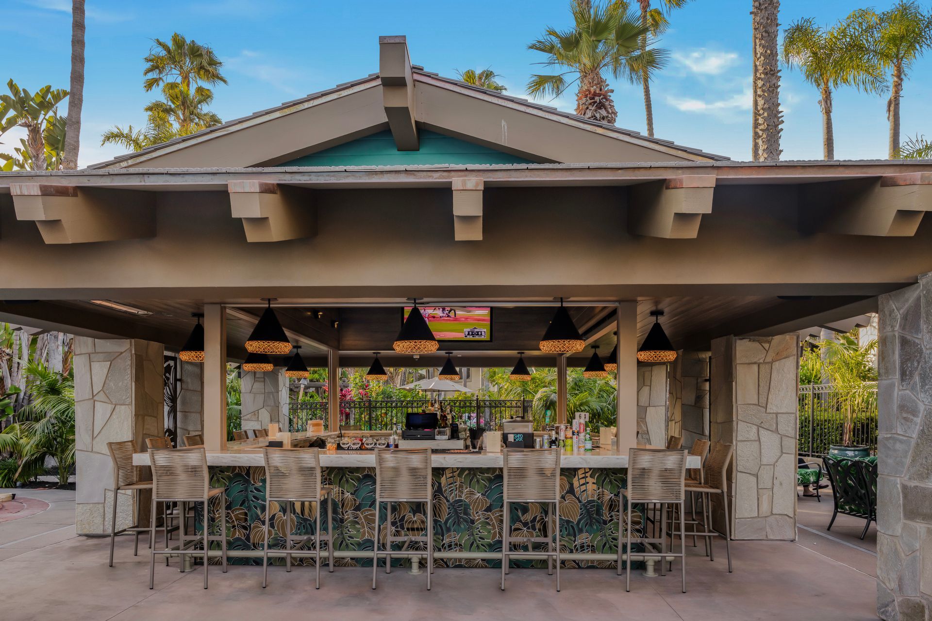 Open-air bar with stools, stone pillars, hanging lights, and palm trees in a tropical courtyard