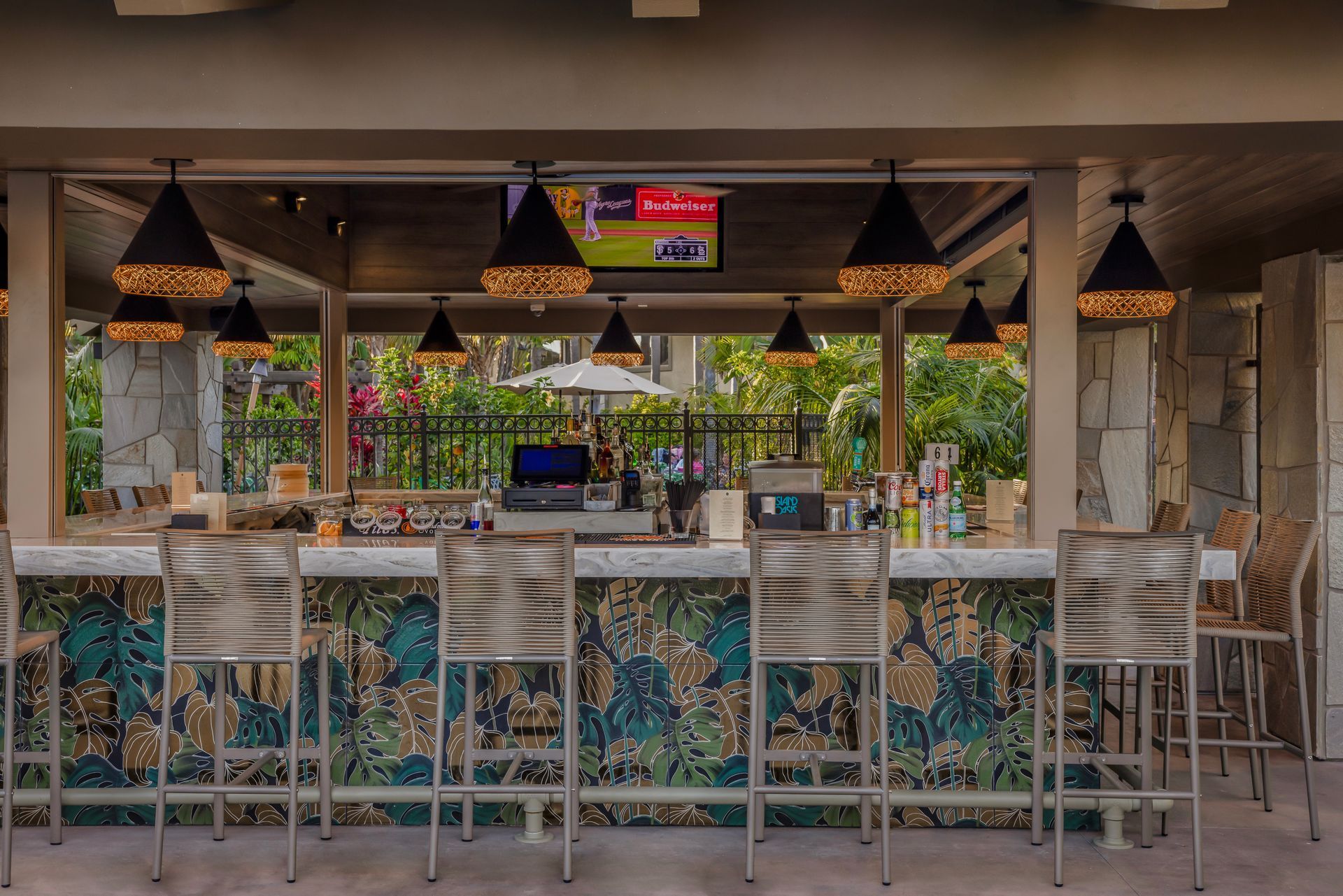 Open-air bar with mosaic counter, white stools, hanging lights, and a small TV screen