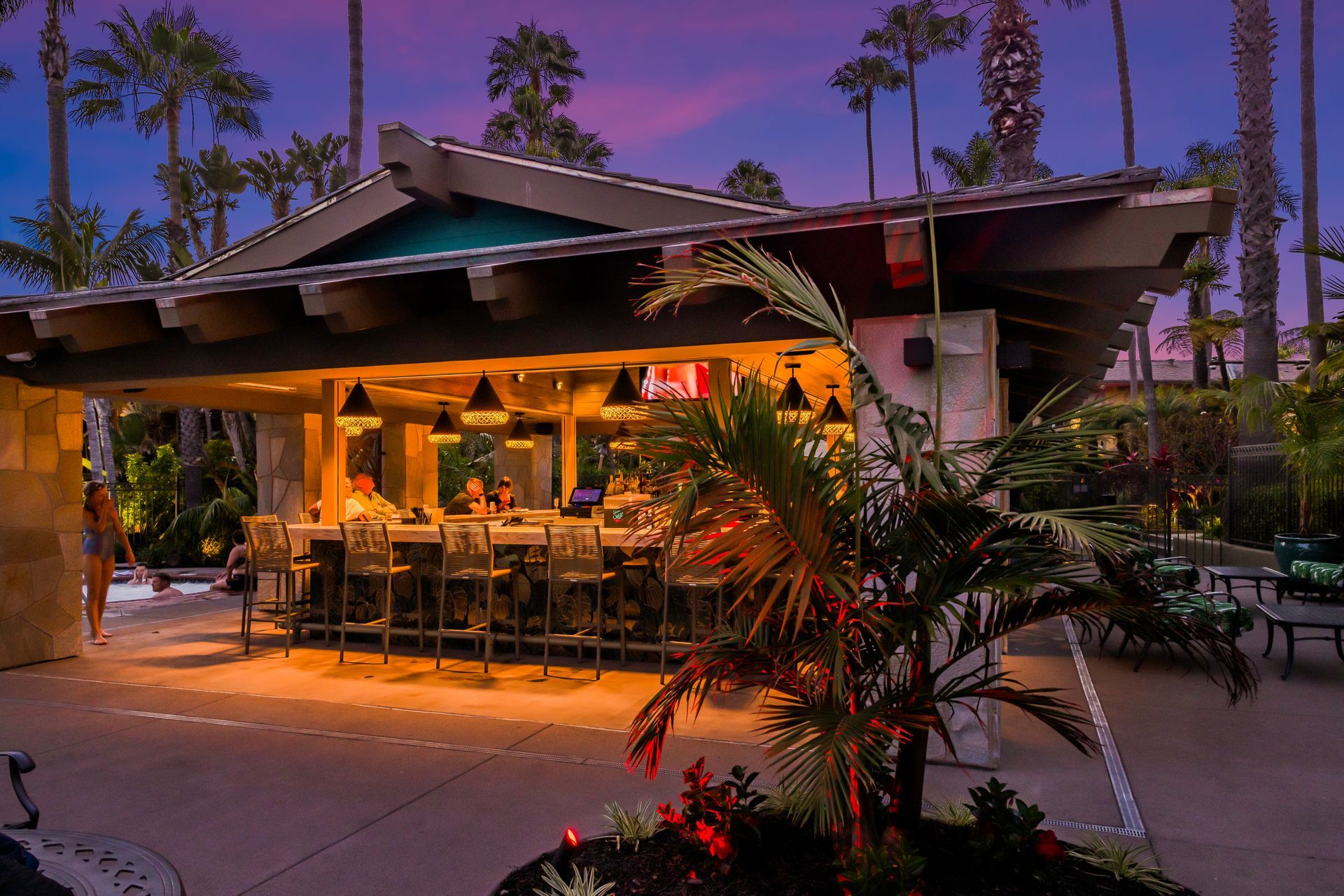 Open-air restaurant at dusk with warm lights, palm trees, and outdoor tables under a purple sky