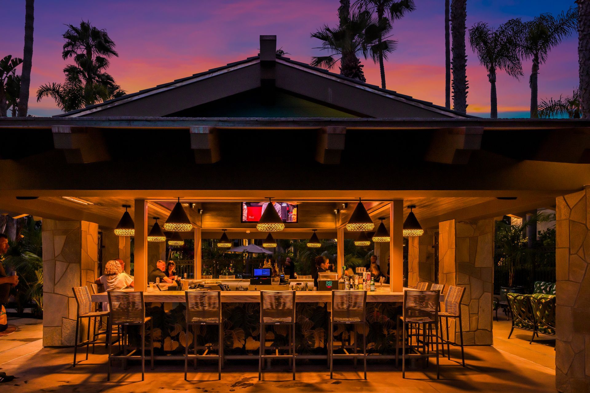 Open-air restaurant at dusk with warm lighting, tables set inside, and palm trees silhouetted against a pink sky