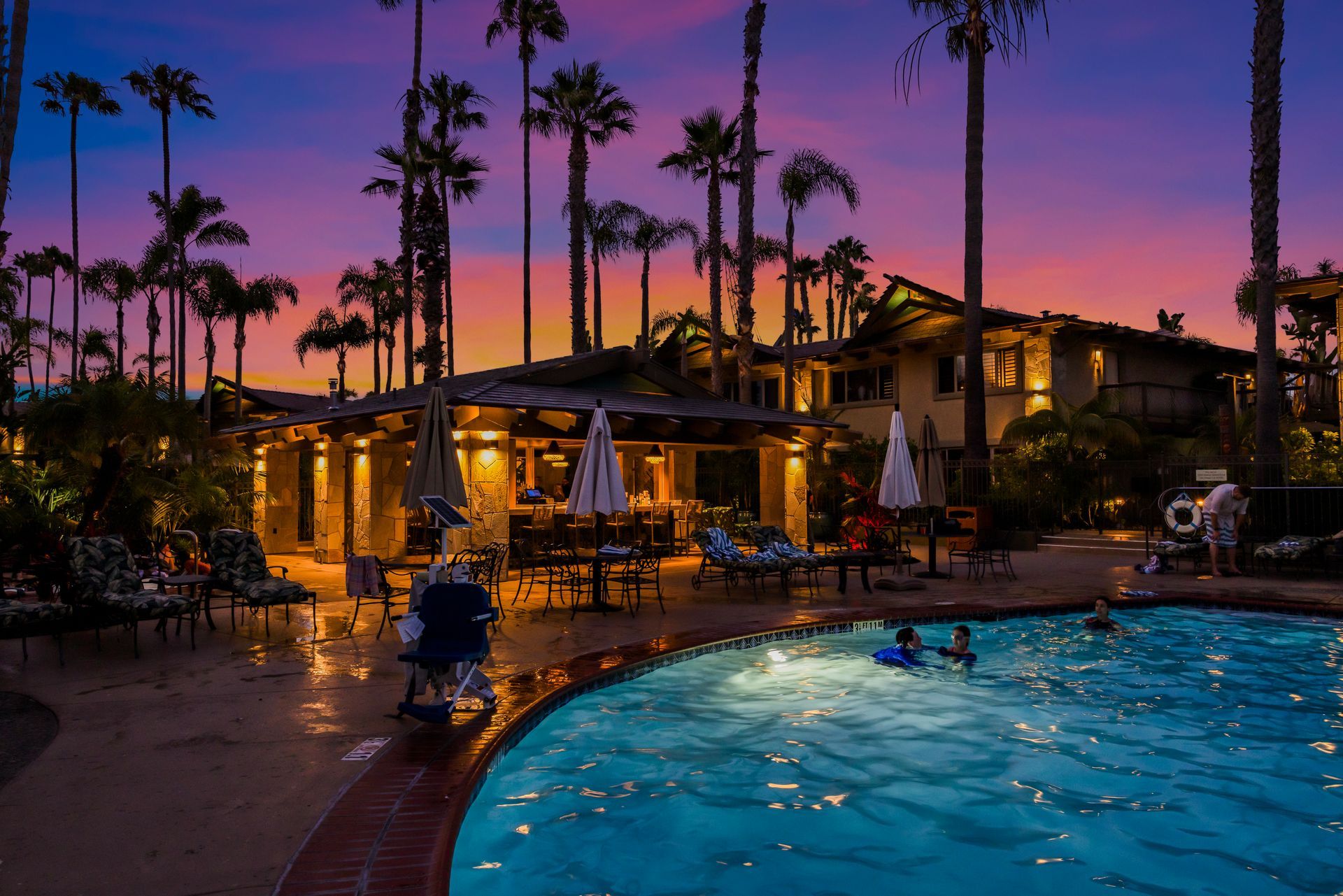 Evening resort pool with palm trees, glowing cabanas, and swimmers under a pink-purple sunset sky